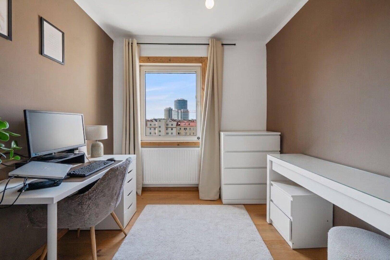 Helles Arbeitszimmer mit Schreibtisch, Computer und Blick auf die Stadt durch das Fenster.