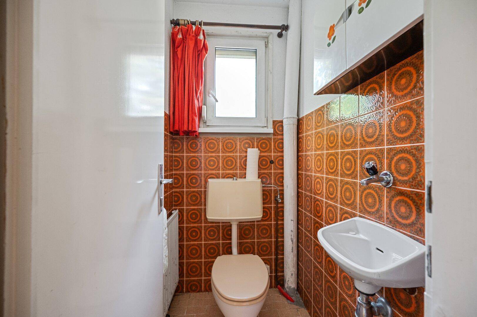 Gäste-WC mit orangefarbenen Fliesen, WC und kleinem Waschbecken im Originalzustand.