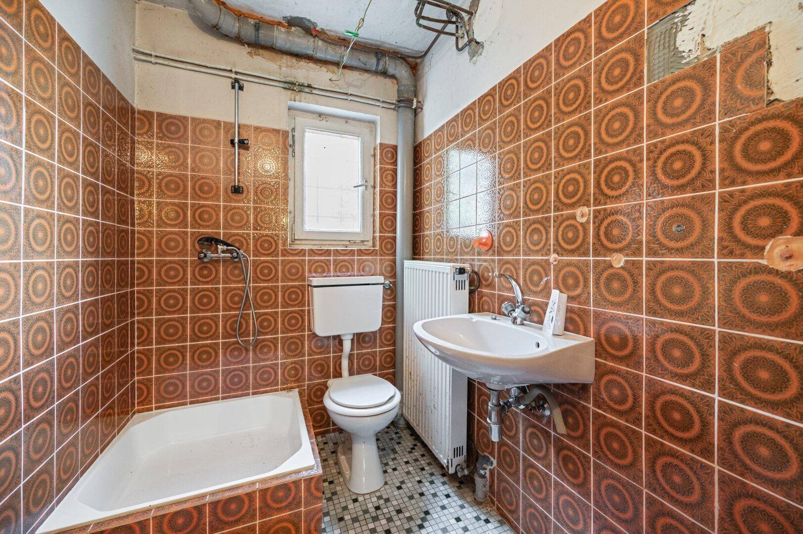 Veraltetes Badezimmer mit braunen Fliesen, Badewanne und einfacher Ausstattung, renovierungsbedürftig.