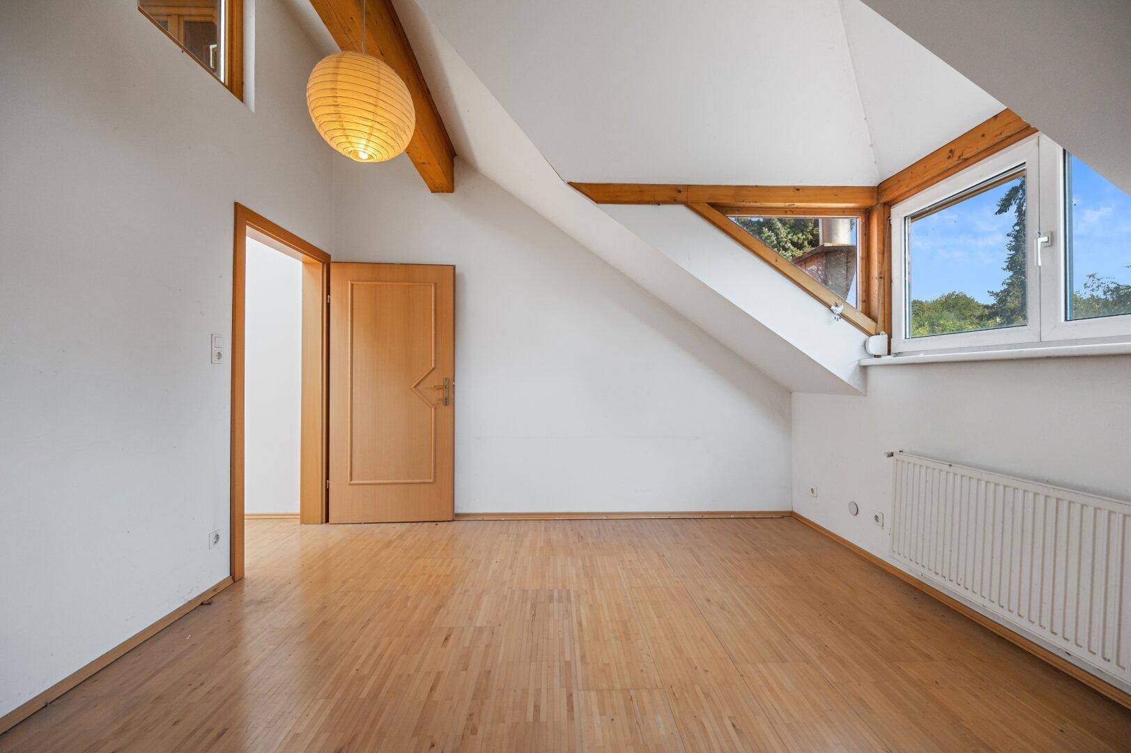 Leerer Raum mit Dachschräge, Holzbalken und großem Fenster, das viel Licht hereinlässt.