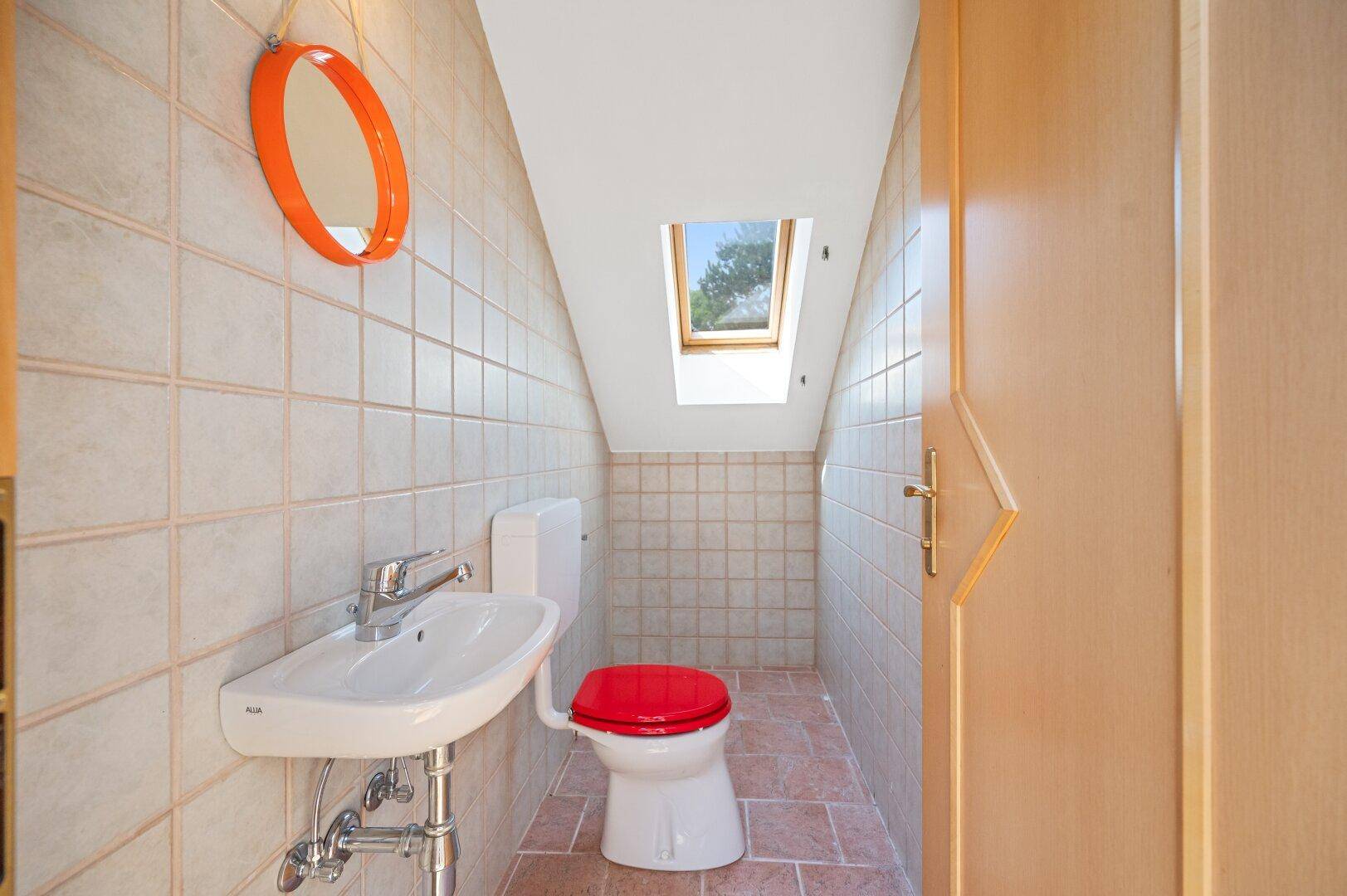 Einfaches Gäste-WC mit Fliesen, Dachfenster und rotem Toilettensitz.