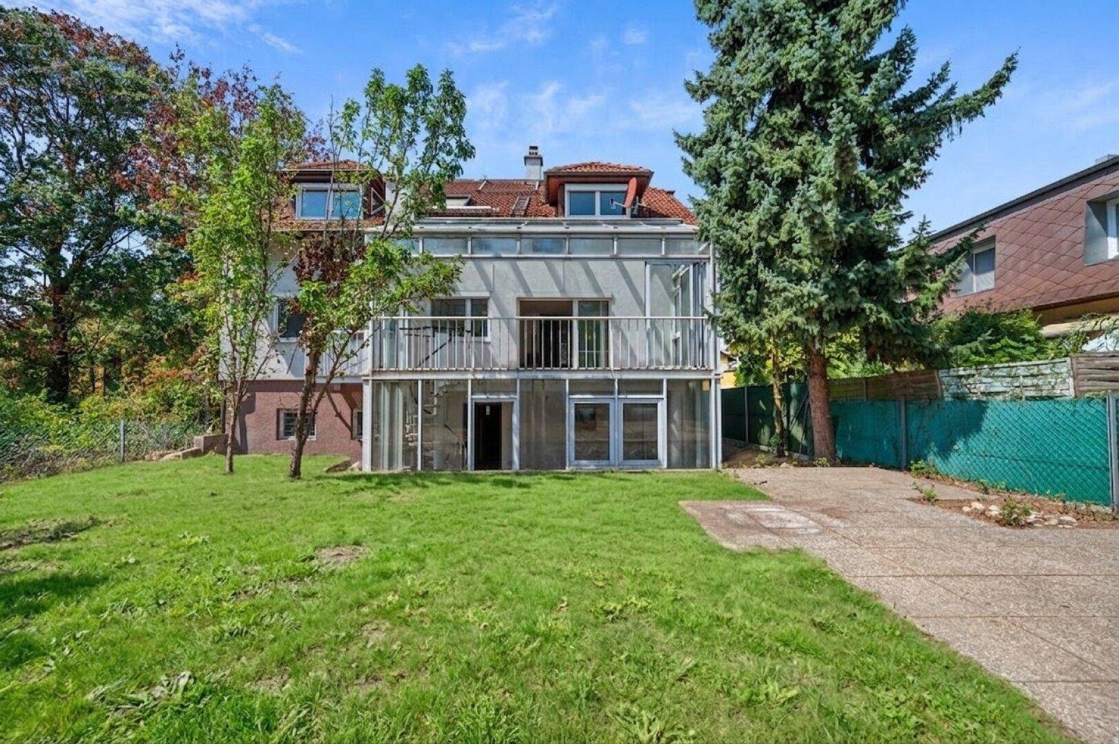 Hinteransicht eines Hauses mit großem Wintergarten, Balkon und weitläufigem grünen Garten.