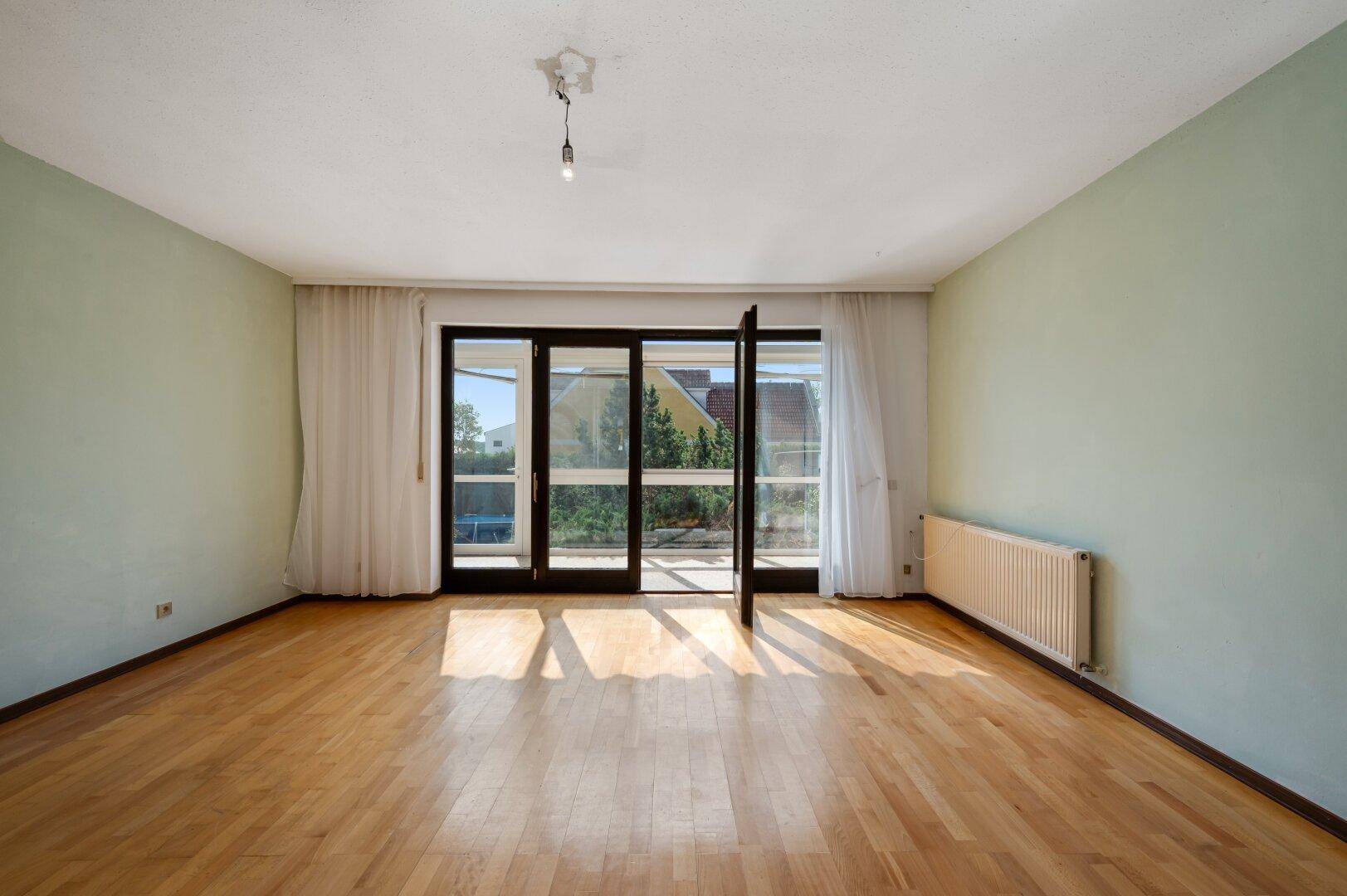 Geräumiges, unmöbliertes Wohnzimmer mit Parkettboden, hellgrünen Wänden und großen Fenstertüren.