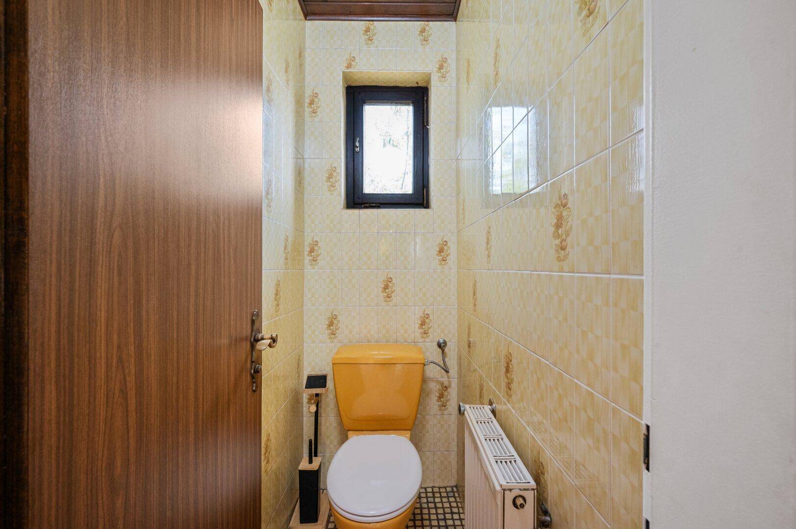 Gäste-WC im Originalzustand mit gelben Fliesen, passender Toilette und einem kleinen Fenster.