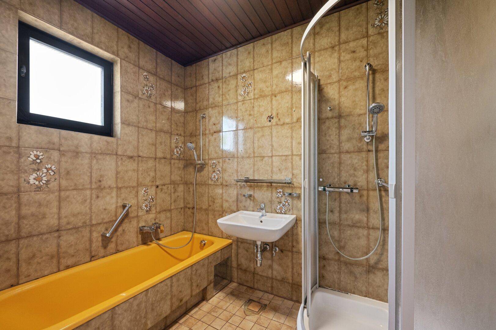 Badezimmer im Originalzustand mit braunen Fliesen, gelber Badewanne und Duschkabine.