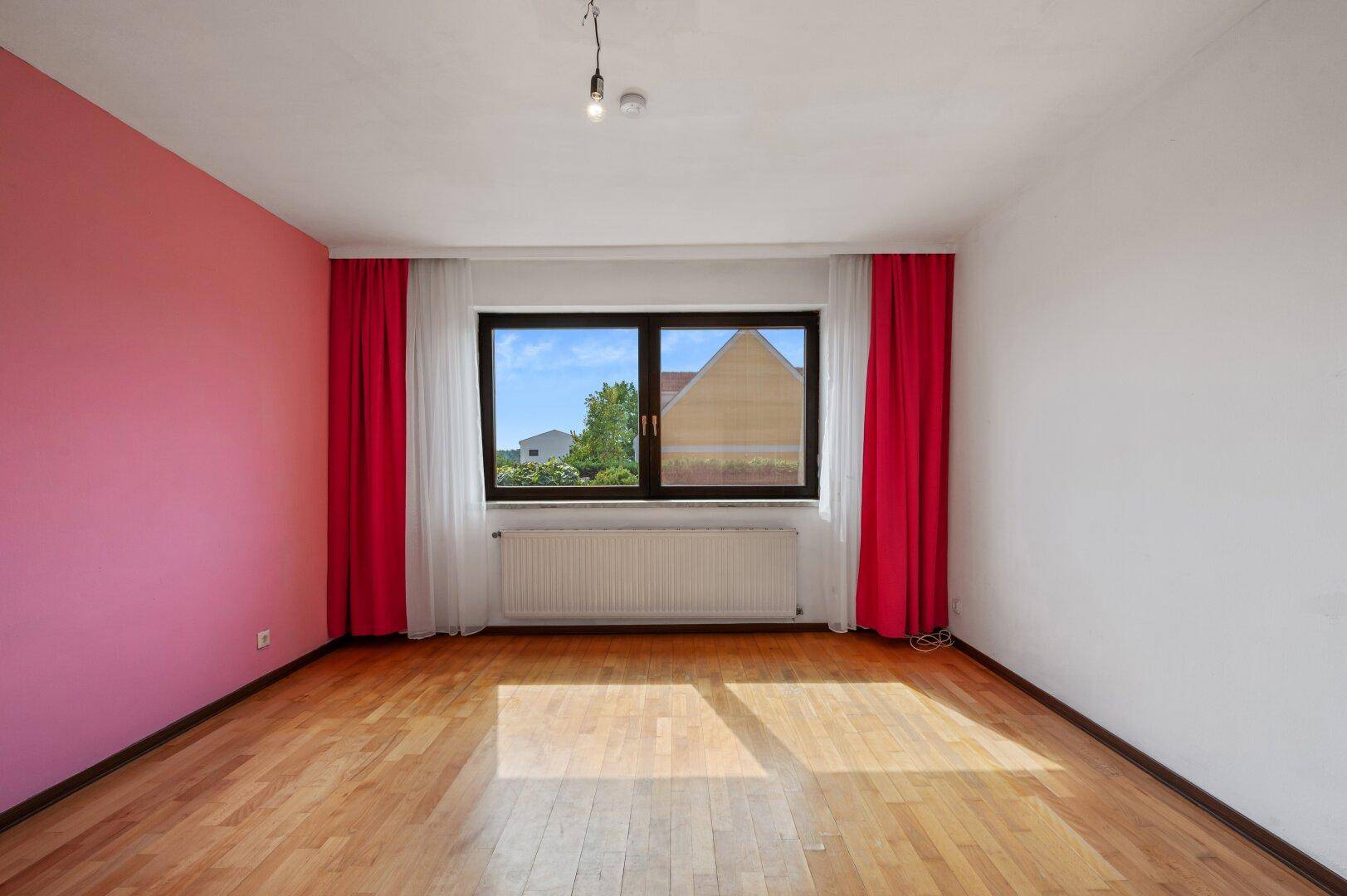 Unmöbliertes Schlafzimmer mit rosa Akzentwand, Parkettboden und einem großen Fenster.