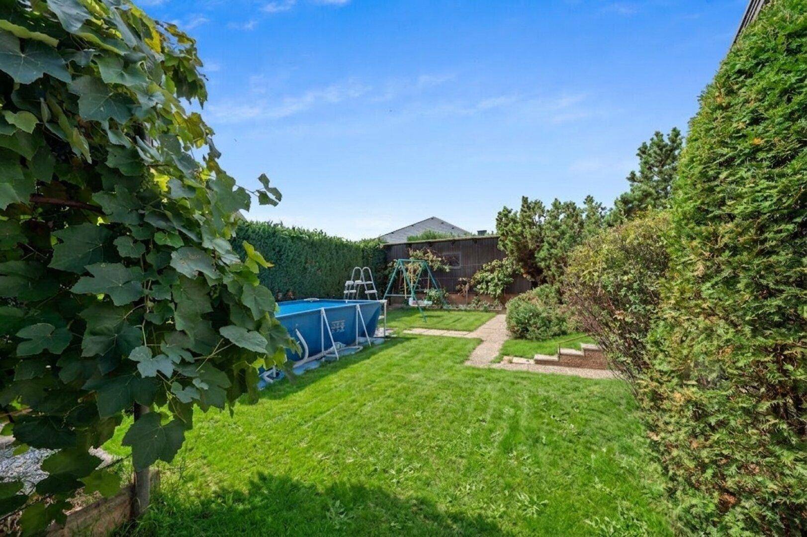 Grüner Garten mit Rasenfläche, Aufstellpool und Kinderschaukel im Hintergrund.