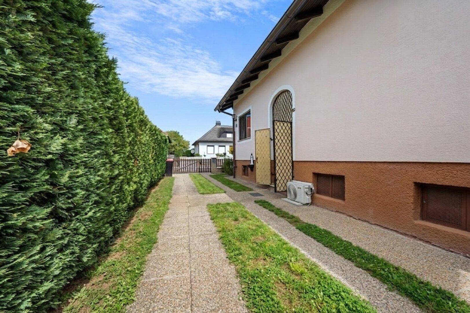 Einfahrt zum Haus mit hoher Hecke und sichtbarer Klimaanlage an der Fassade.