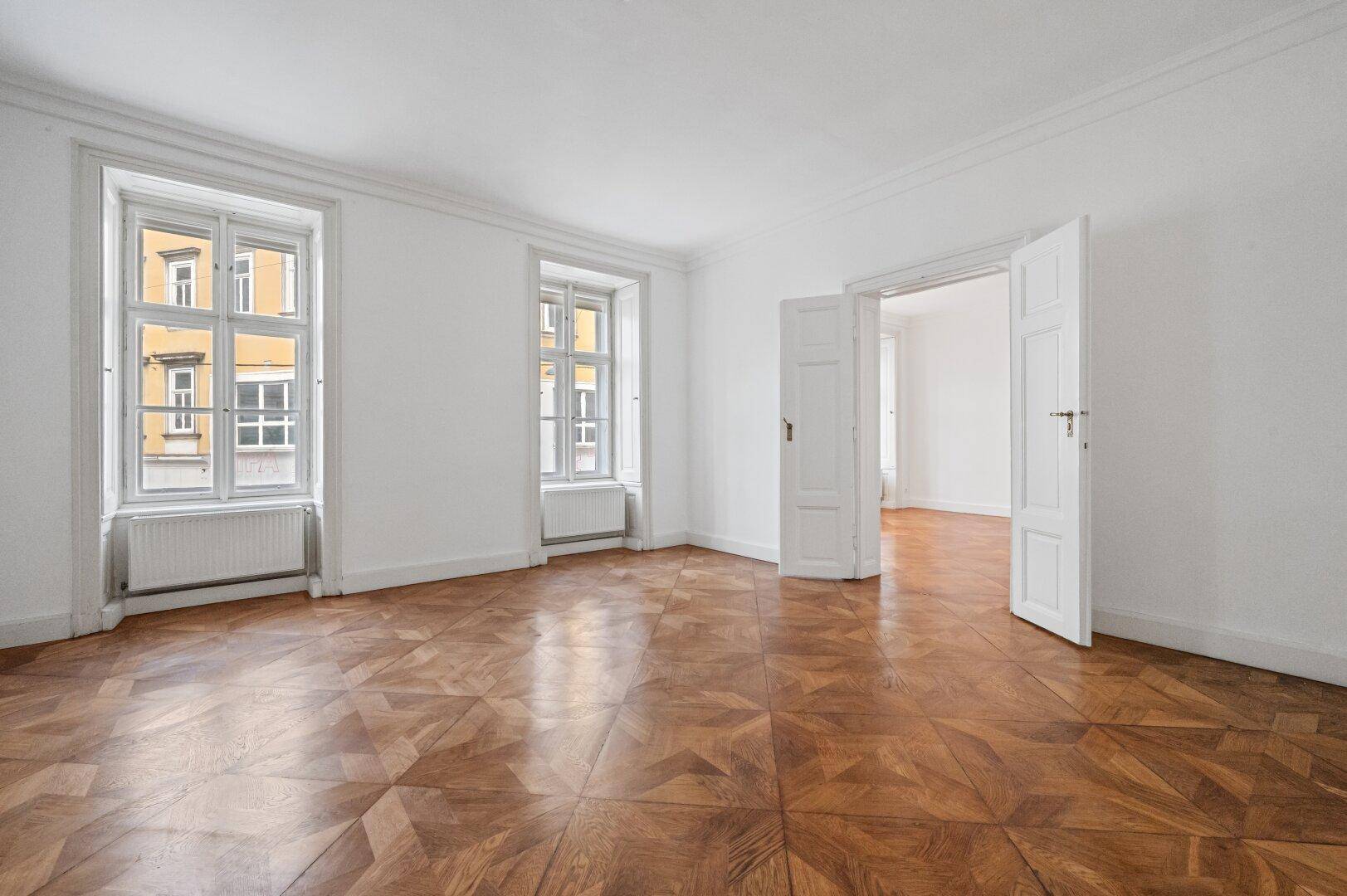 Heller, leerer Raum mit klassischem Parkettboden, hohen Decken und zwei großen Fenstern.