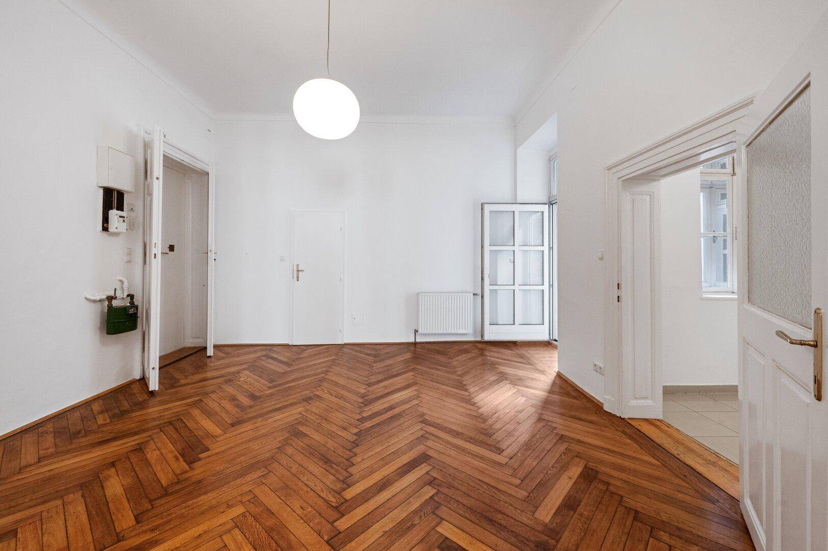 Leerer Raum mit Fischgrätparkett, hohen Decken und einem Fenster, das zu einem Balkon führt.