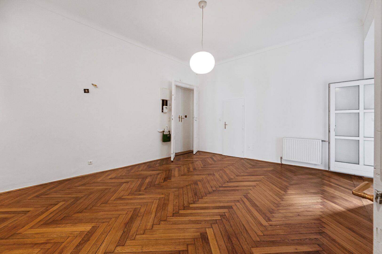 Heller, leerer Raum mit Fischgrätparkett, hohen Decken und einem Fenster, das zu einem Balkon führt.