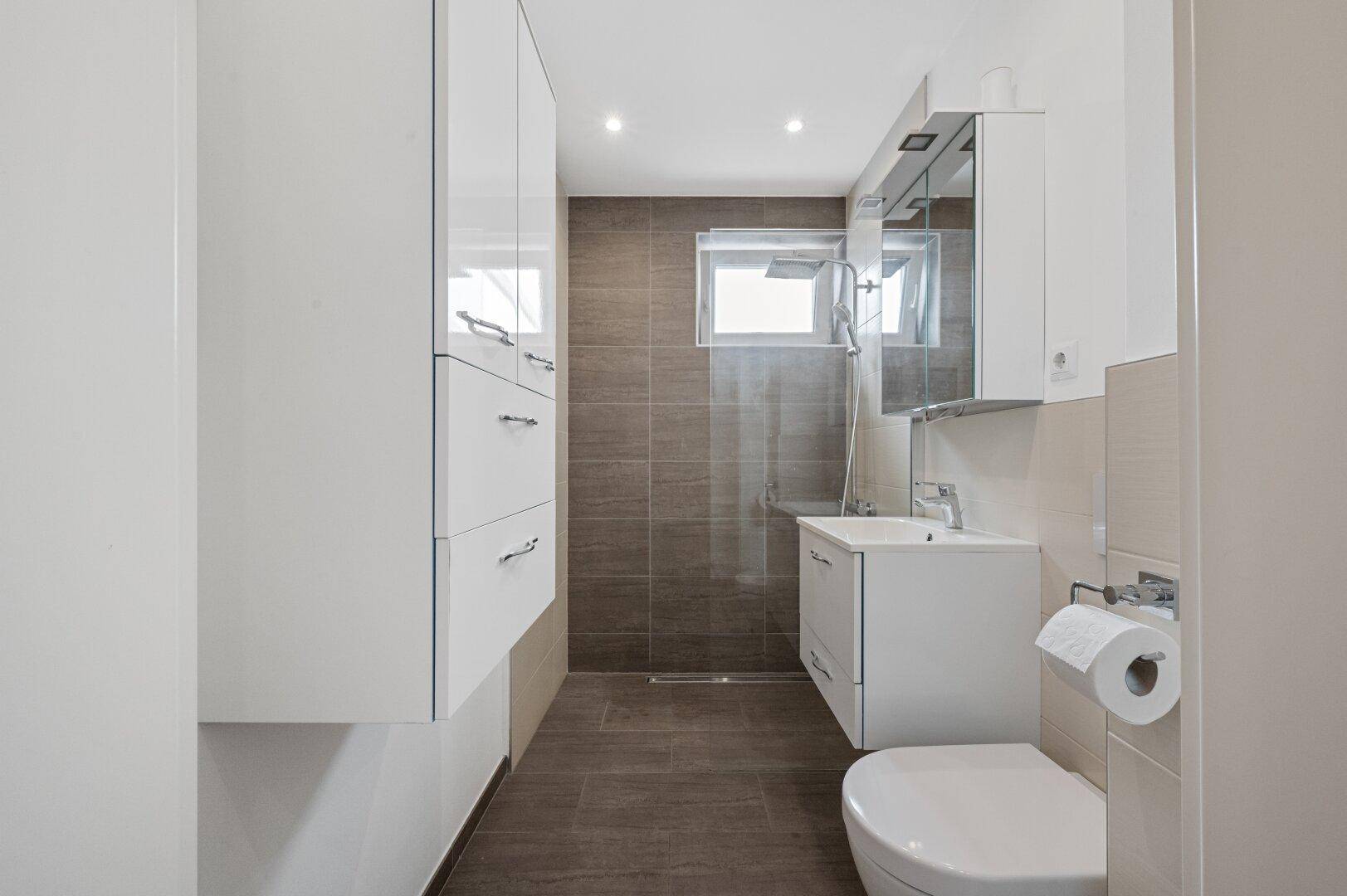 Modernes Badezimmer mit begehbarer Dusche, weißen Schränken und großformatigen Fliesen.
