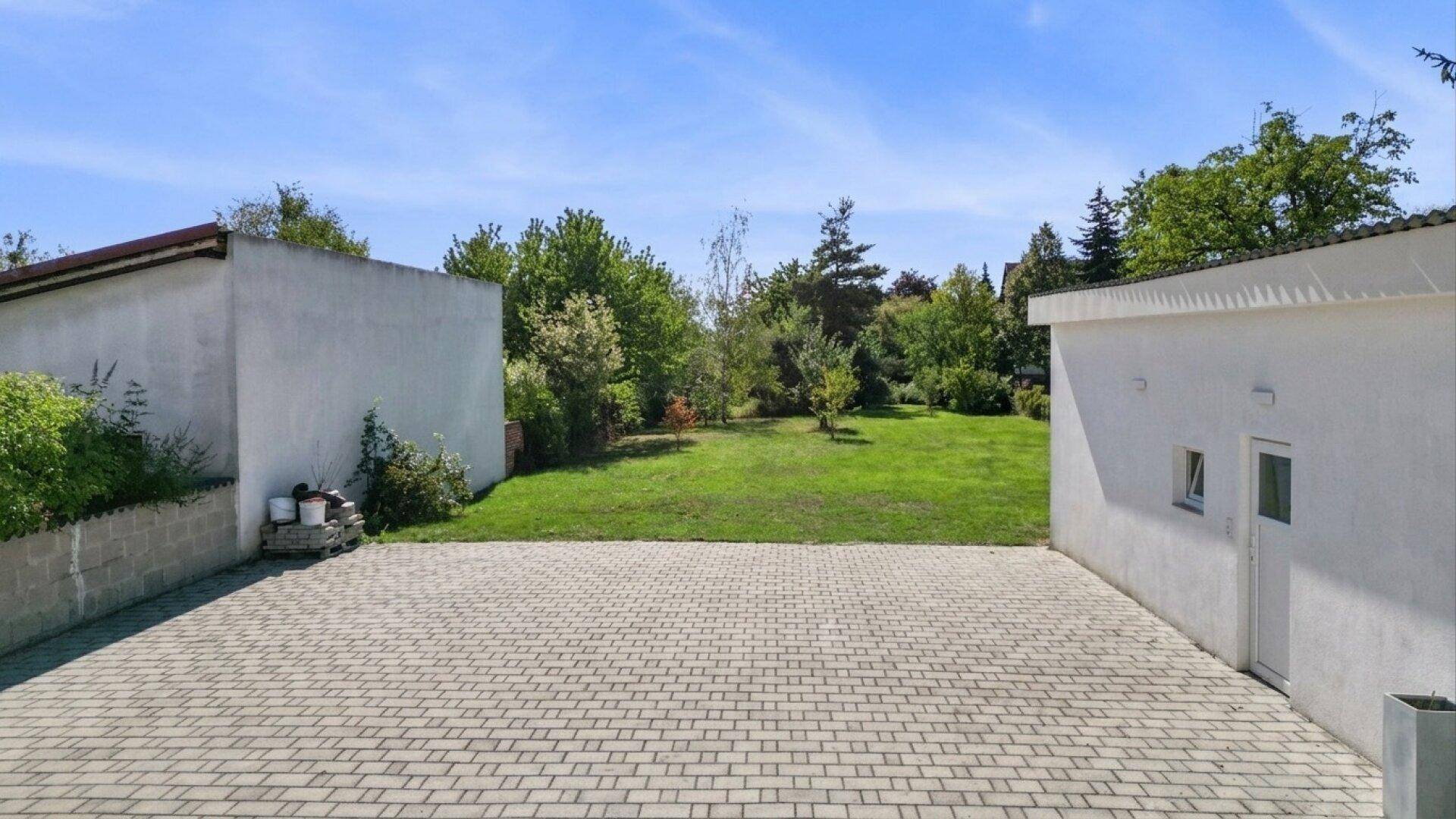 Großer gepflasterter Bereich vor einer Garage, der zu einem weitläufigen grünen Garten führt.