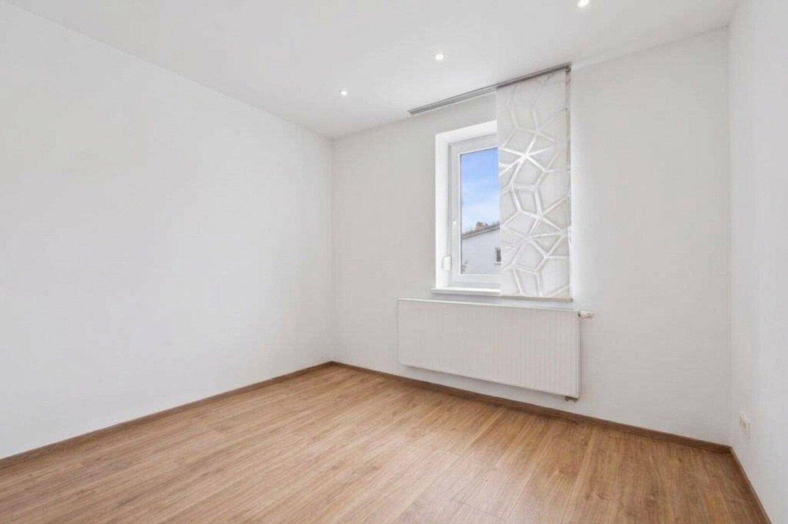 Helles Schlafzimmer mit Laminatboden und großem Fenster, das viel Tageslicht hereinlässt.