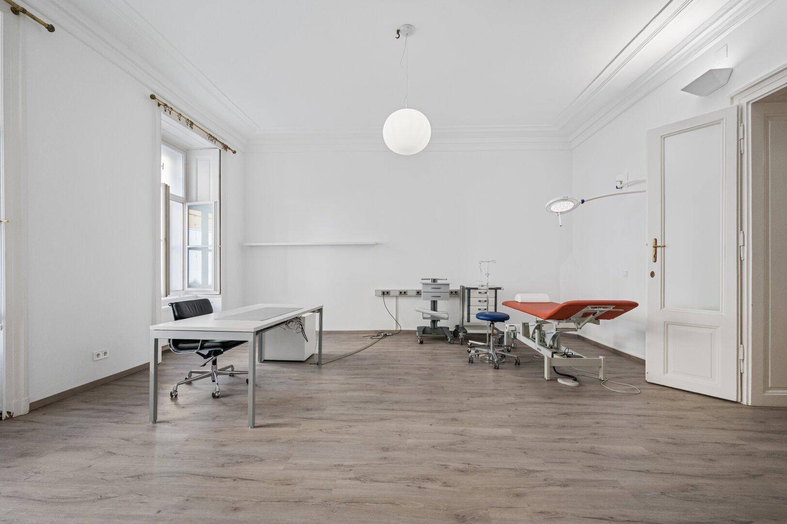 Heller Ordinationsraum mit medizinischer Liege und Schreibtisch, ausgestattet für professionelle Nutzung.