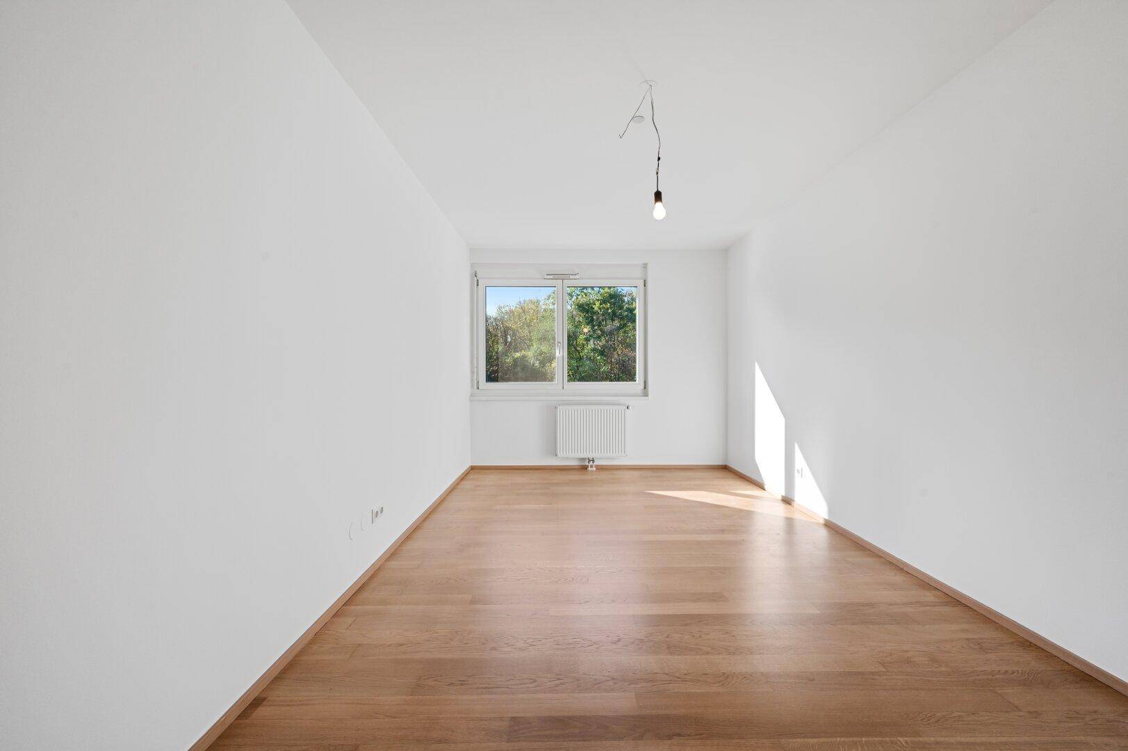 Leeres Schlafzimmer mit Parkettboden und Fenster mit Blick ins Grüne.