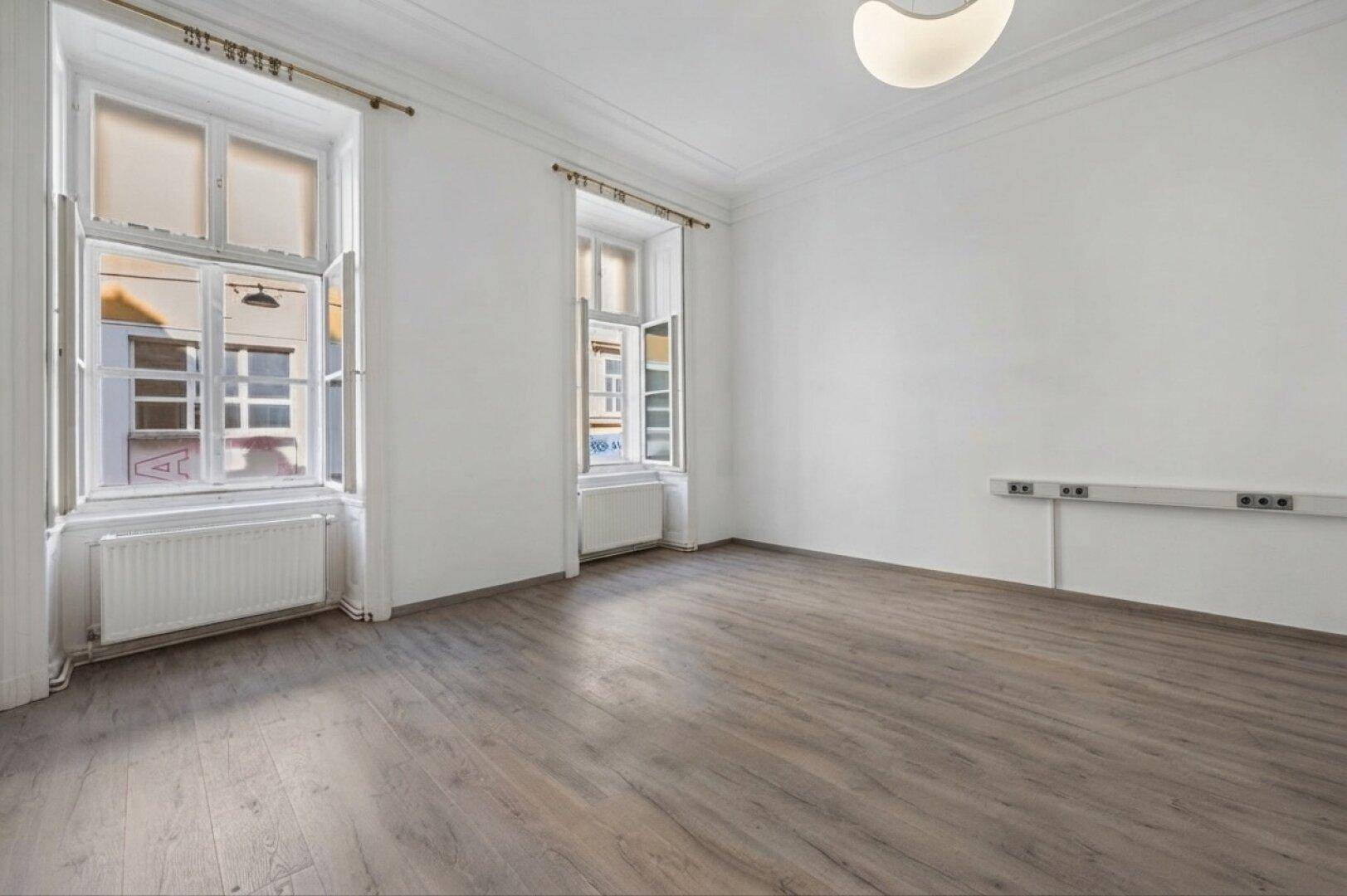 Heller, leerer Raum mit Laminatboden, hohen Decken und zwei großen Fenstern mit Heizkörpern.