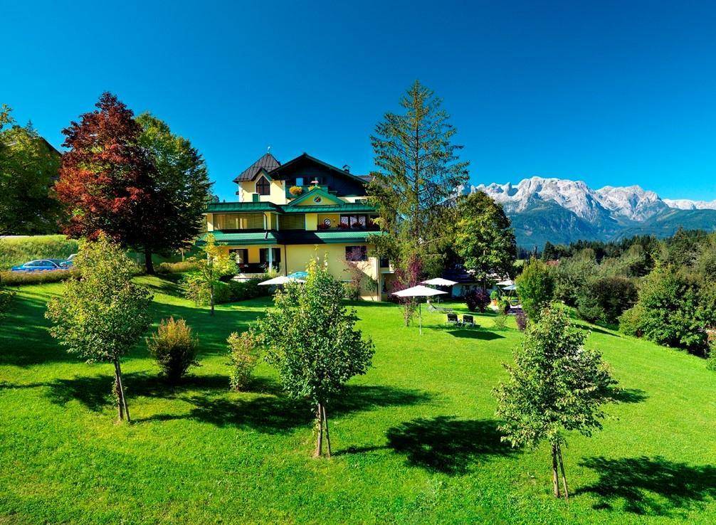 Panoramablick auf das Gebäude inmitten grüner Hügel und Bäume mit majestätischen Bergen im Hintergrund.