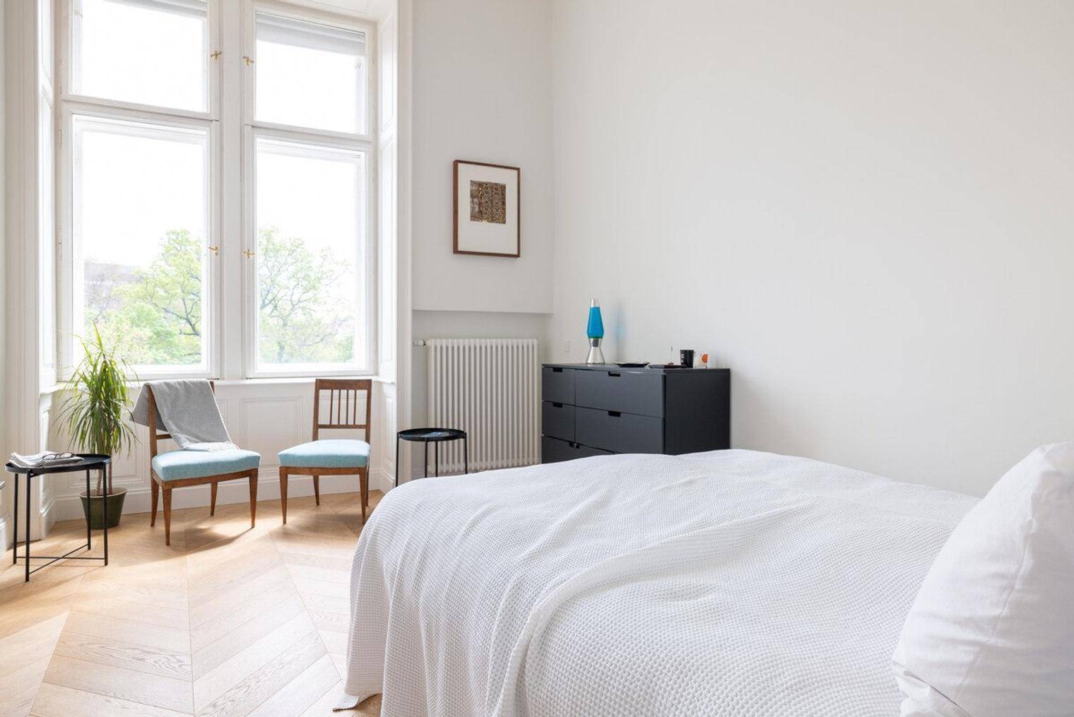 Helles Schlafzimmer mit großen Fenstern, Fischgrätparkett und einem bequemen Bett.