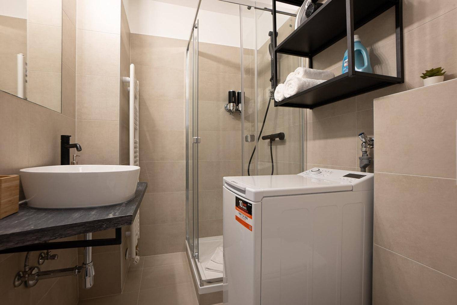 Modernes Badezimmer mit Dusche, Waschbecken und integrierter Waschmaschine in beigen Fliesen.