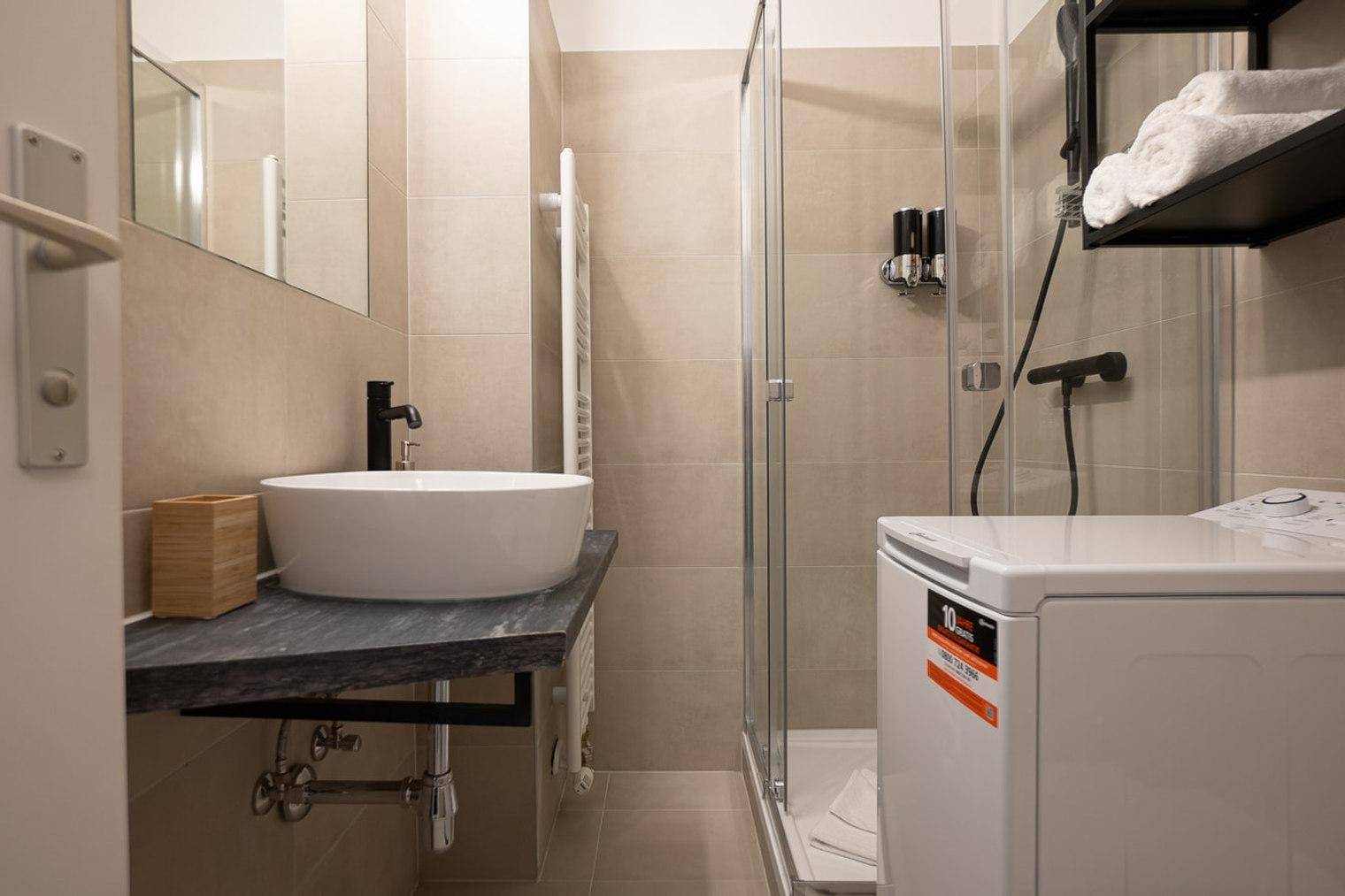 Modernes Badezimmer mit Dusche, Waschbecken und integrierter Waschmaschine in beigen Fliesen.
