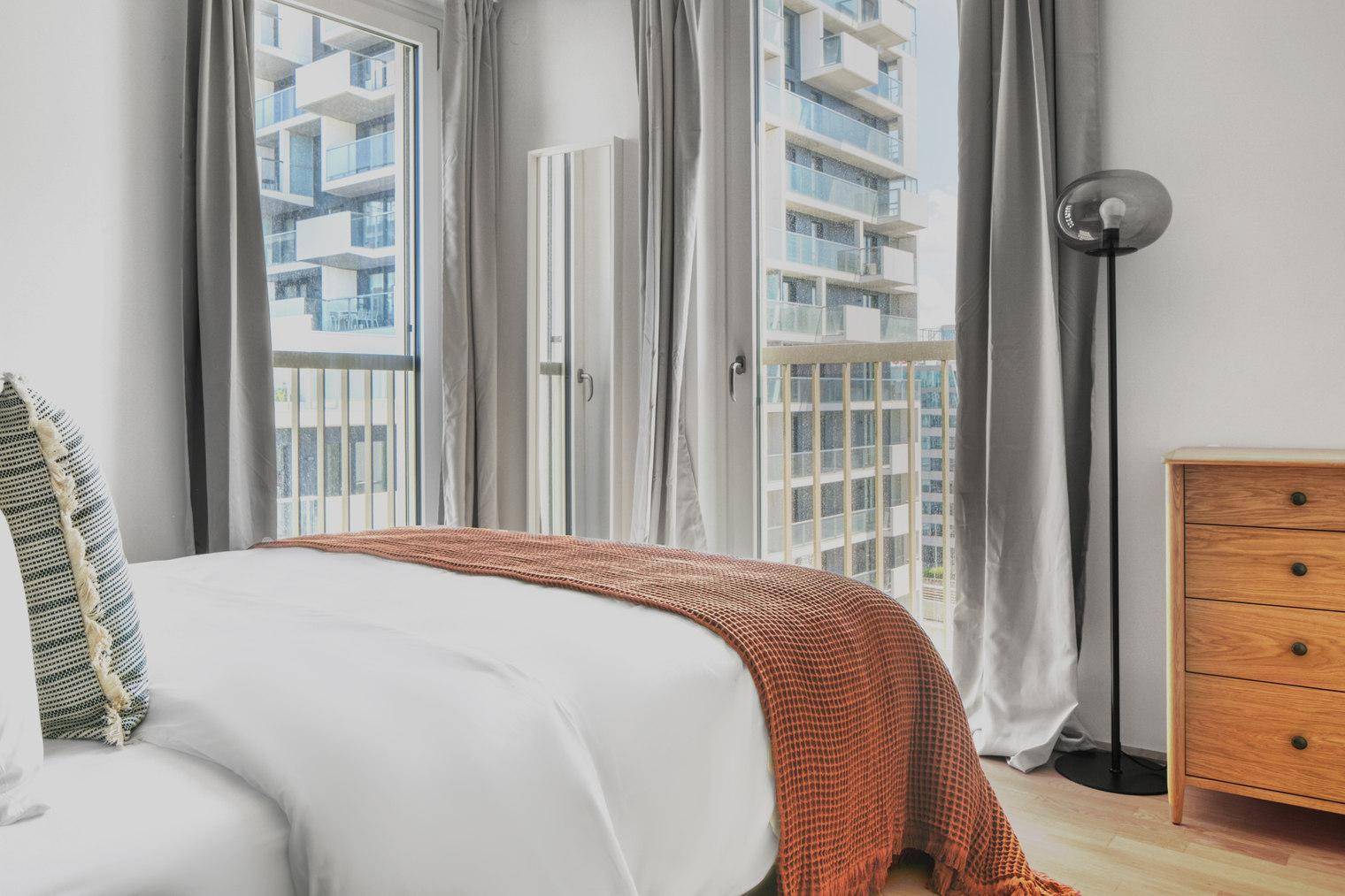 Schlafzimmer mit Doppelbett, Kommode und großen Fenstern mit Stadtblick.