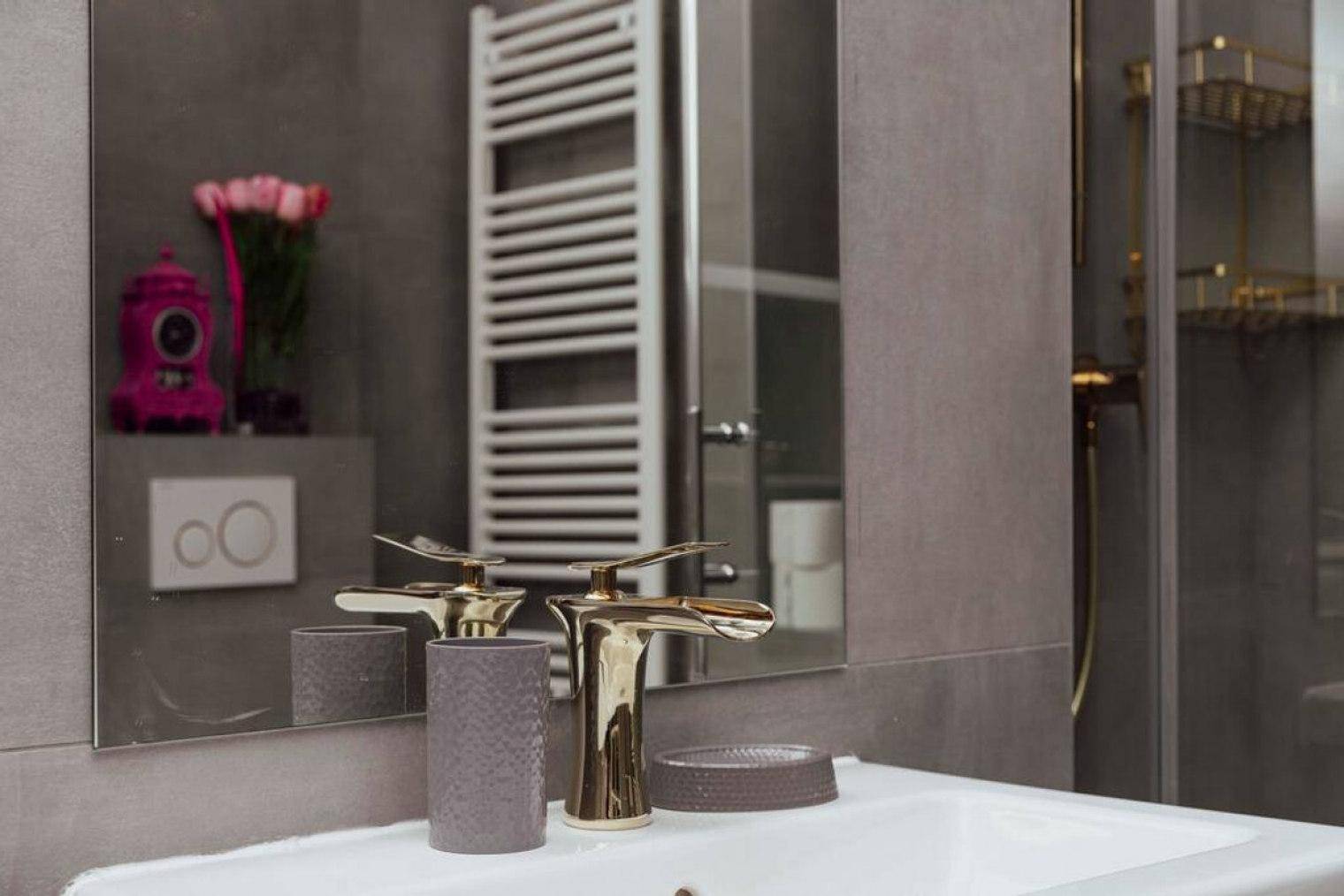 Modernes Badezimmer mit grauen Fliesen, elegantem goldenem Wasserhahn und großem Spiegel über dem Waschbecken.