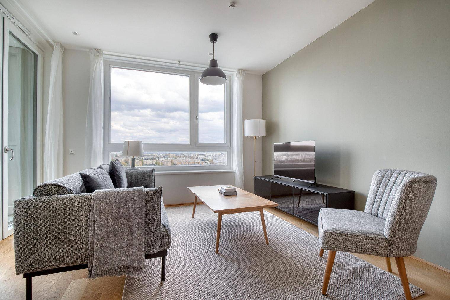 Gemütliches Wohnzimmer mit Sofa, Sessel und Blick auf die Stadt durch große Fenster.