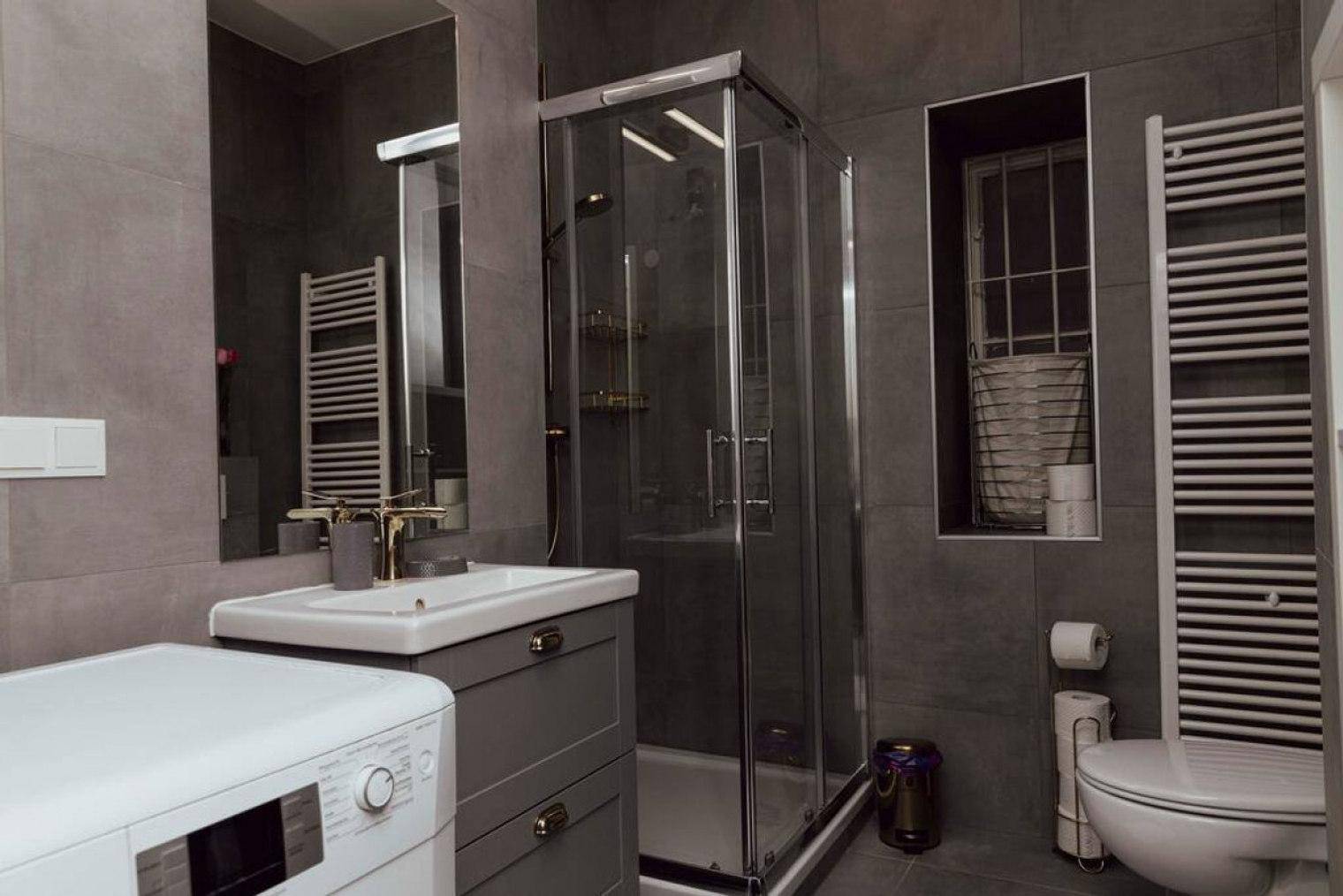 Modernes Badezimmer mit grauen Fliesen, Duschkabine, Waschbecken, Toilette und Waschmaschine.