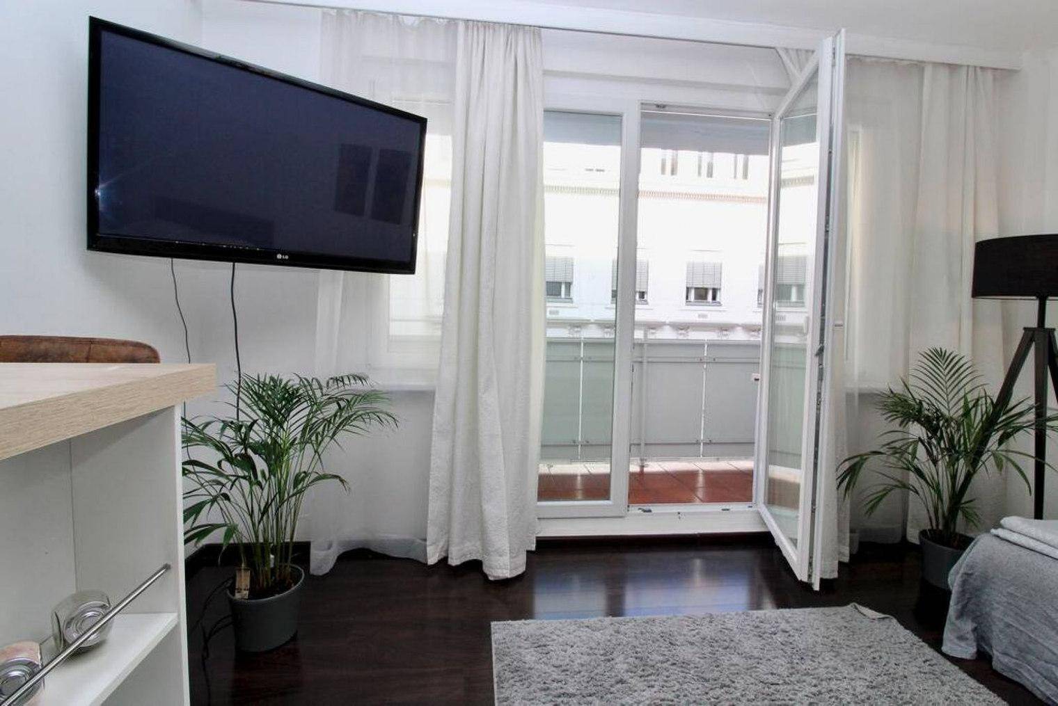 Heller Wohnbereich mit Fernseher, Pflanzen und geöffneten Balkontüren.