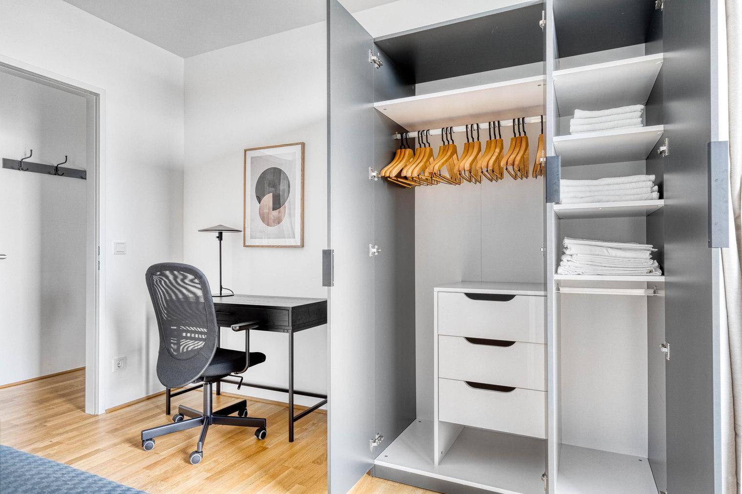 Schlafzimmer mit Schreibtisch, Bürostuhl und offenem Kleiderschrank.