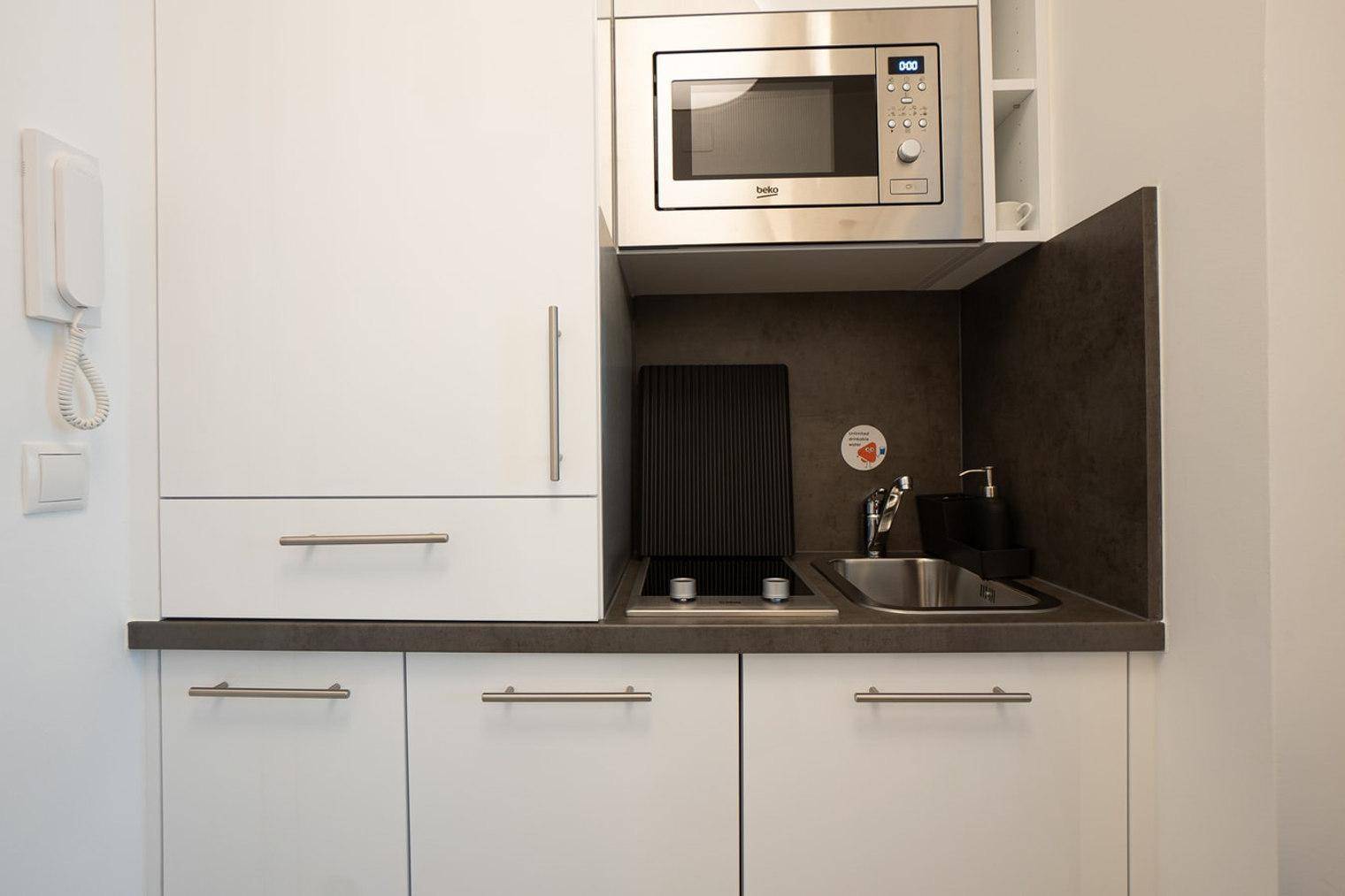 Kompakte Küchenzeile mit Mikrowelle, Kochfeld und Spüle in modernem, weißem Design.