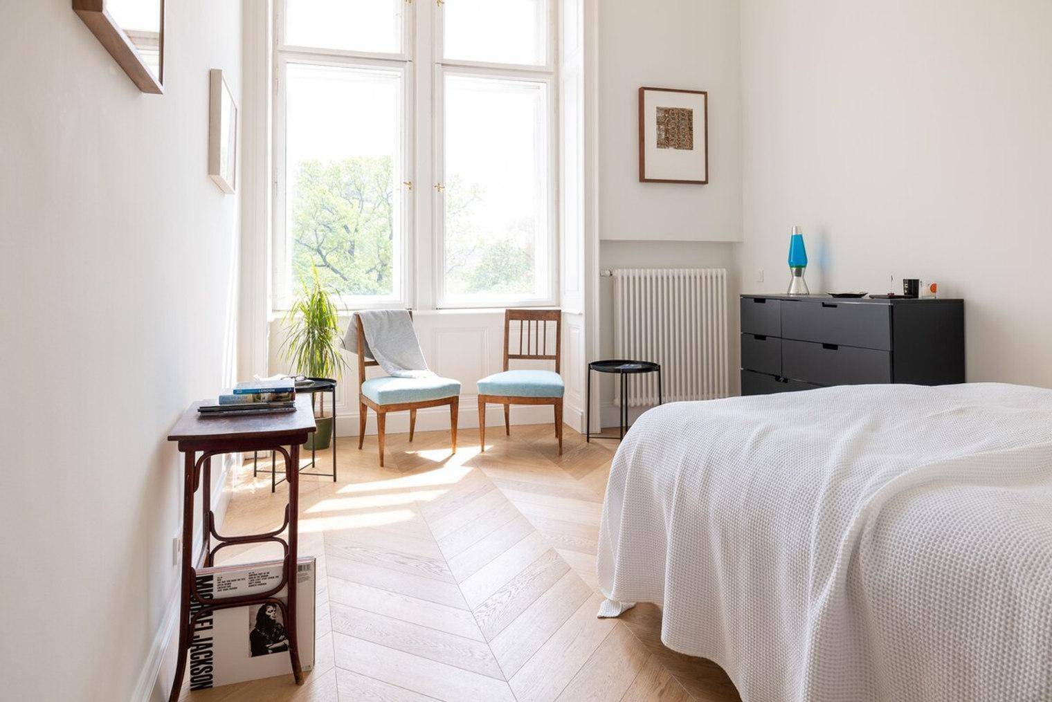 Helles Schlafzimmer mit Fischgrätparkett, hohen Fenstern und einem modernen Sideboard.