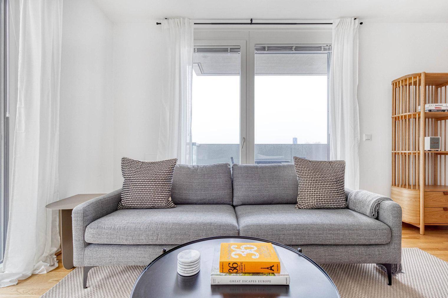 Gemütliches Wohnzimmer mit grauem Sofa, Couchtisch und großen Fenstern.