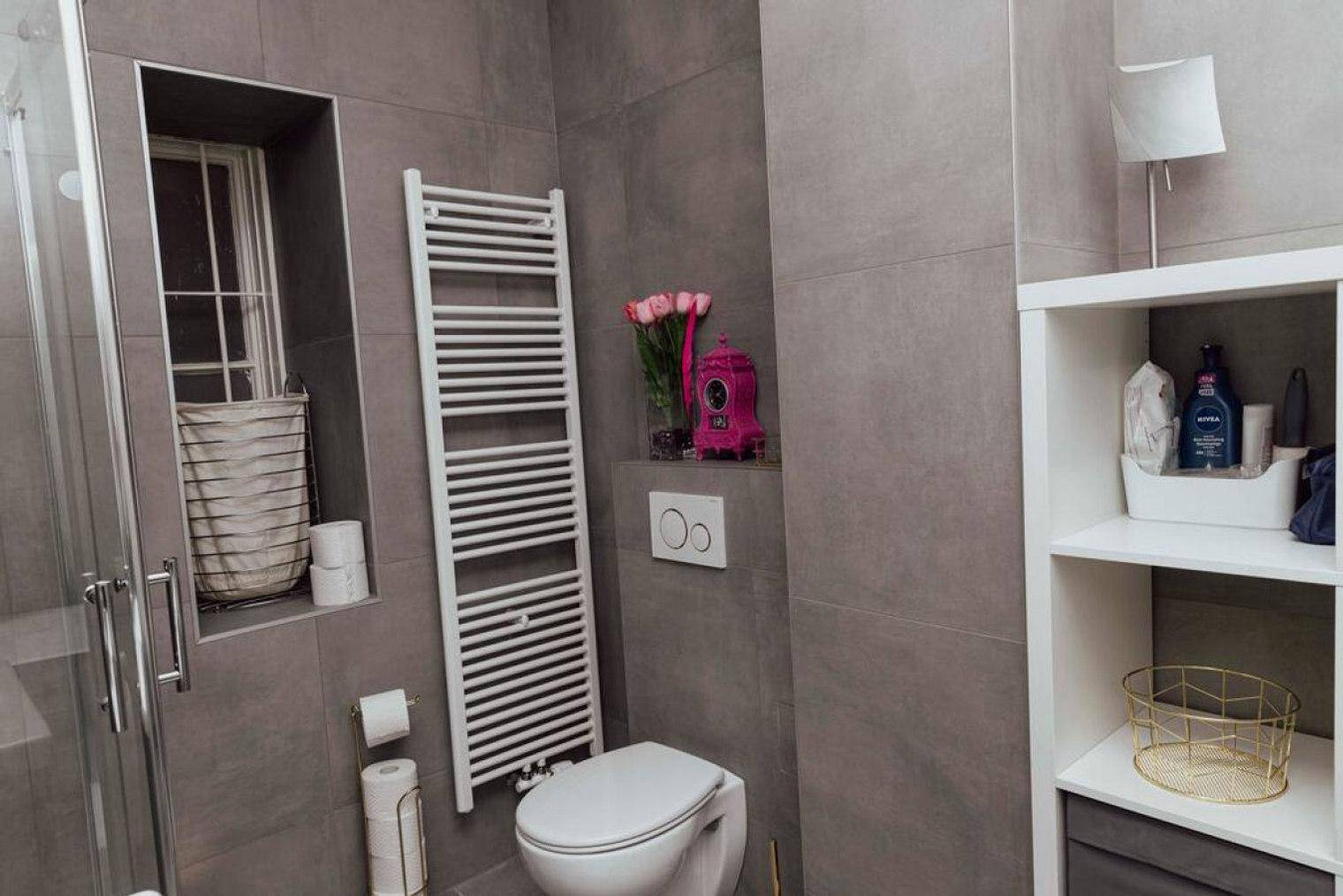 Modernes Badezimmer mit grauen Fliesen, Toilette, beheiztem Handtuchhalter und praktischem Regal.