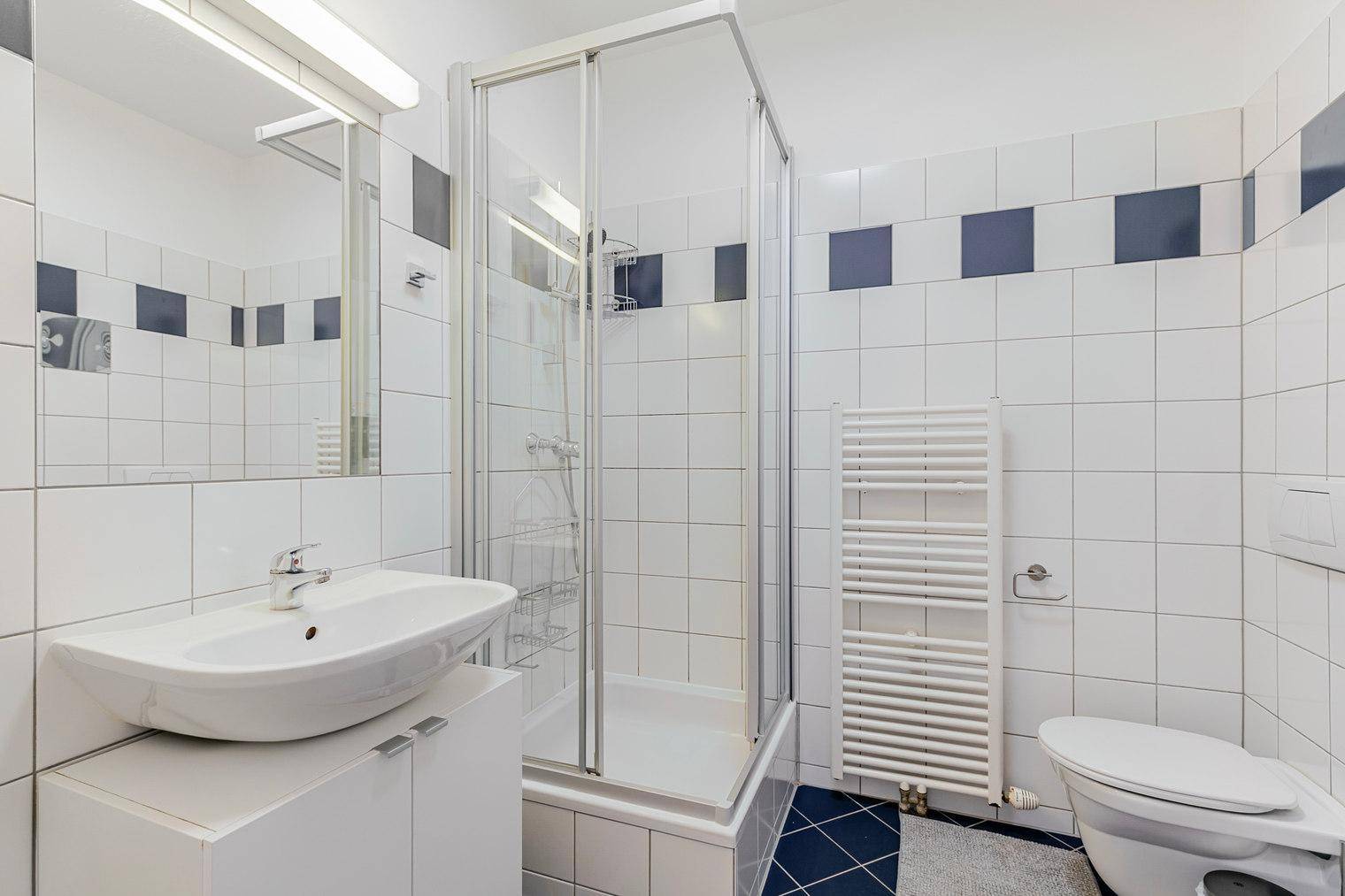 Badezimmer mit Duschkabine, Waschbecken und Toilette, gefliest in Weiß und Dunkelblau.
