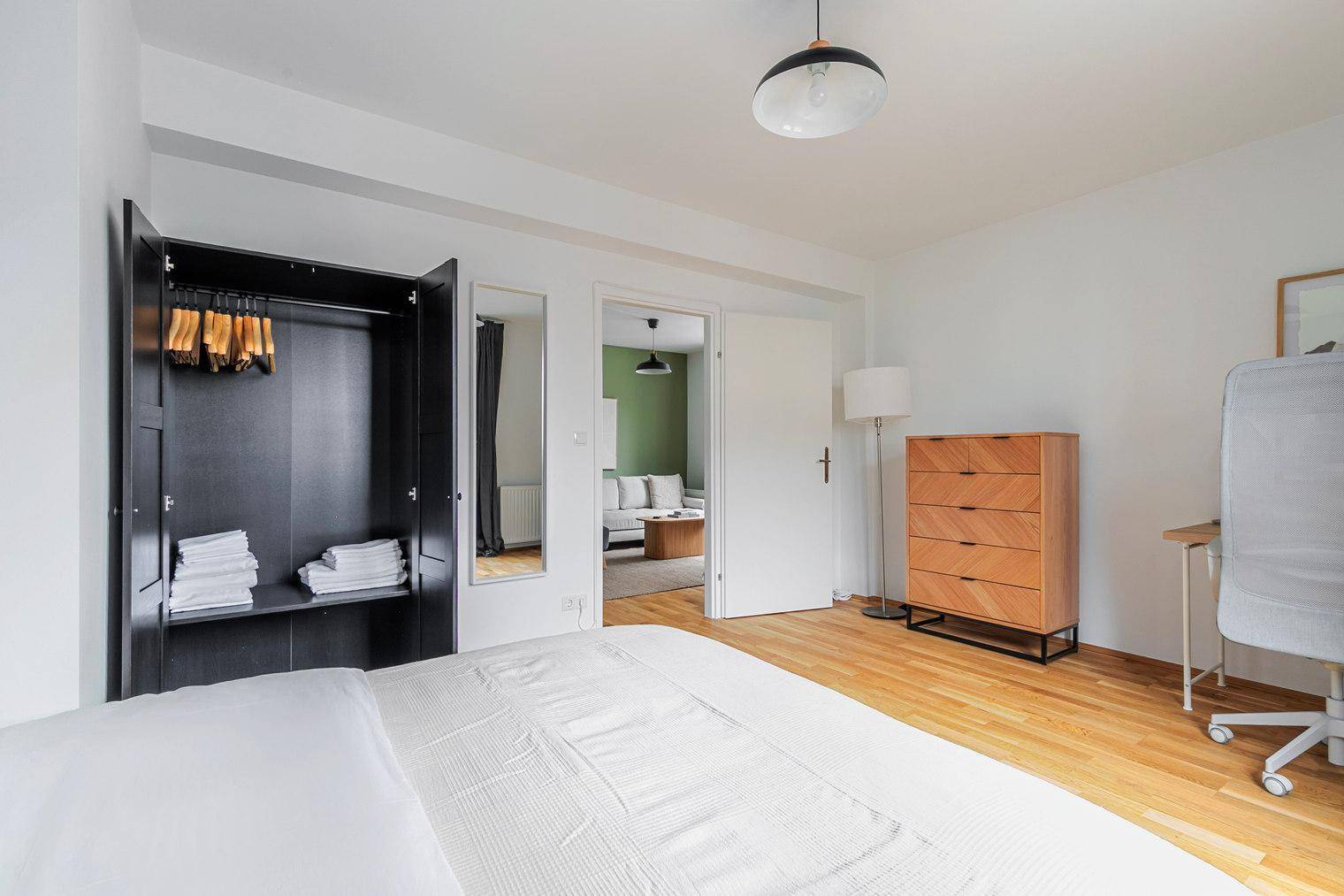 Schlafzimmer mit Kleiderschrank, Schreibtisch und Blick durch die offene Tür zum Wohnzimmer.