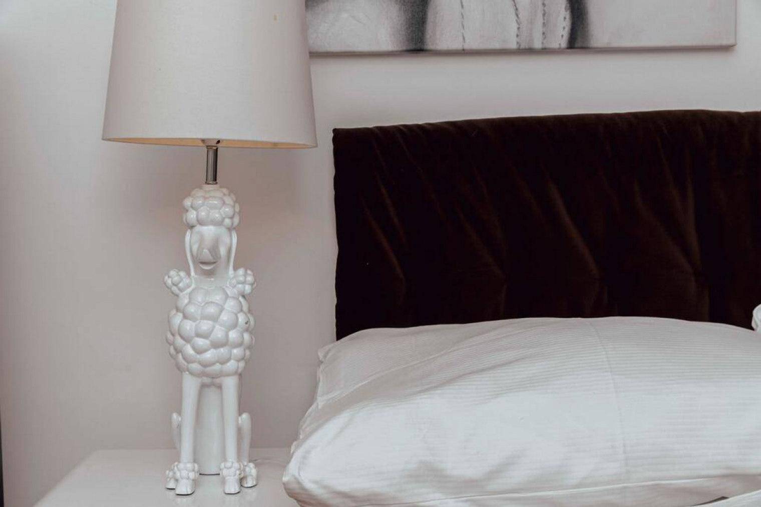 Detailansicht einer Nachttischlampe in Form eines Pudels neben einem Bett im Schlafzimmer.