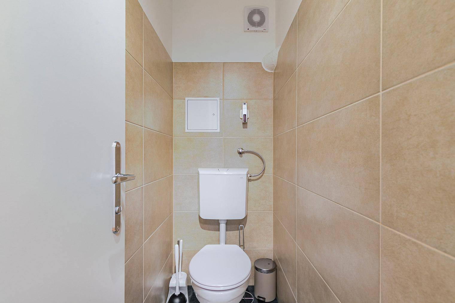 Separates WC mit Toilette, Fliesen an den Wänden und einem Abluftventilator an der Decke.