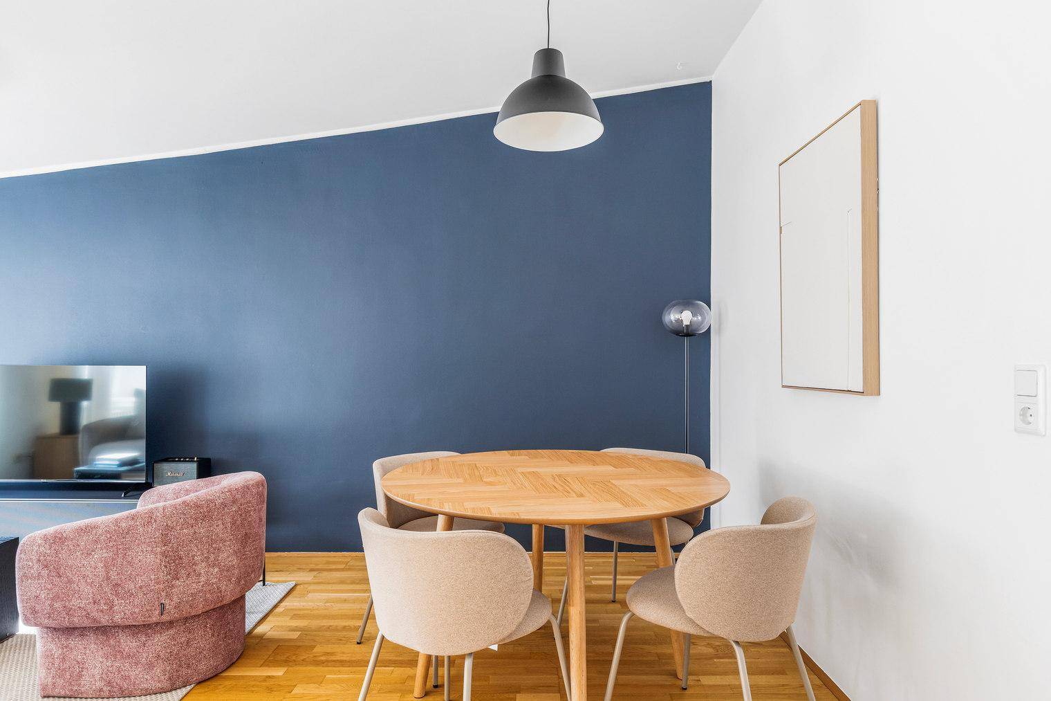 Moderner Essbereich mit rundem Holztisch und bequemen Stühlen vor einer blauen Akzentwand.