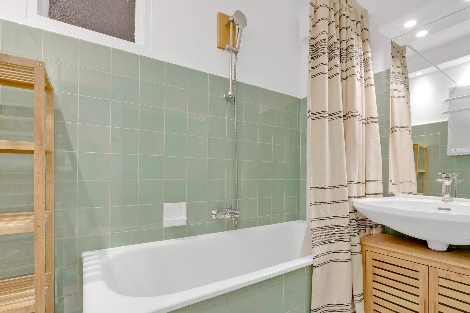 Badezimmer mit Badewanne, grünen Fliesen und einem Waschbecken mit Holzunterschrank.