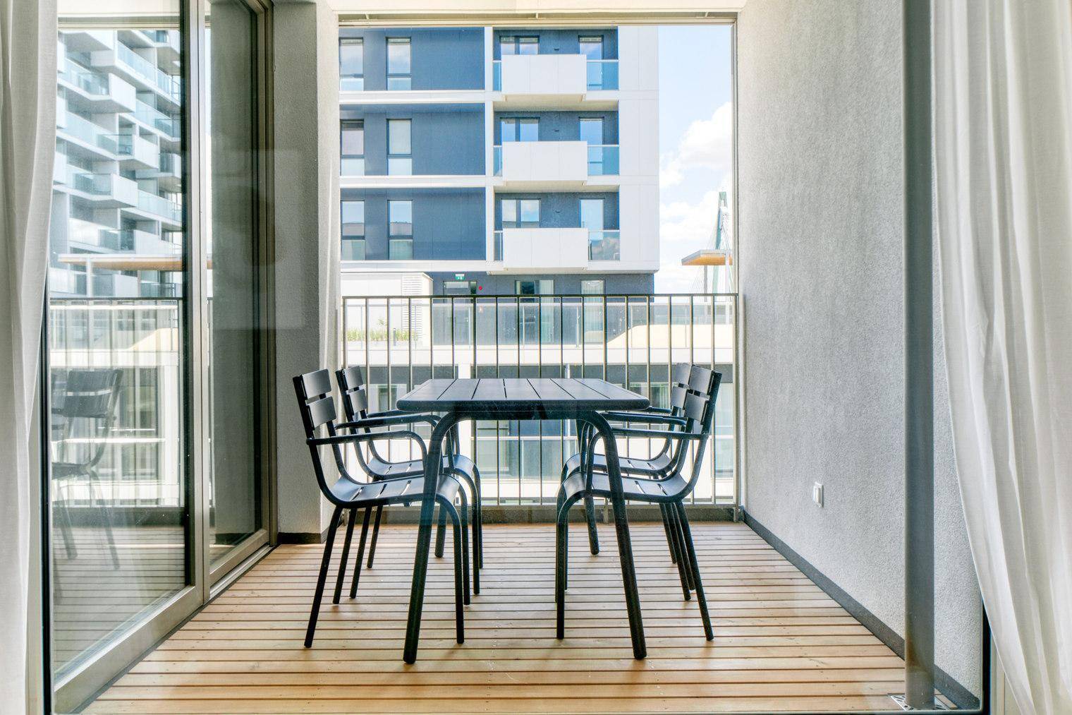 Geräumiger Balkon mit Holzboden und Esstisch, umgeben von modernen Gebäuden.