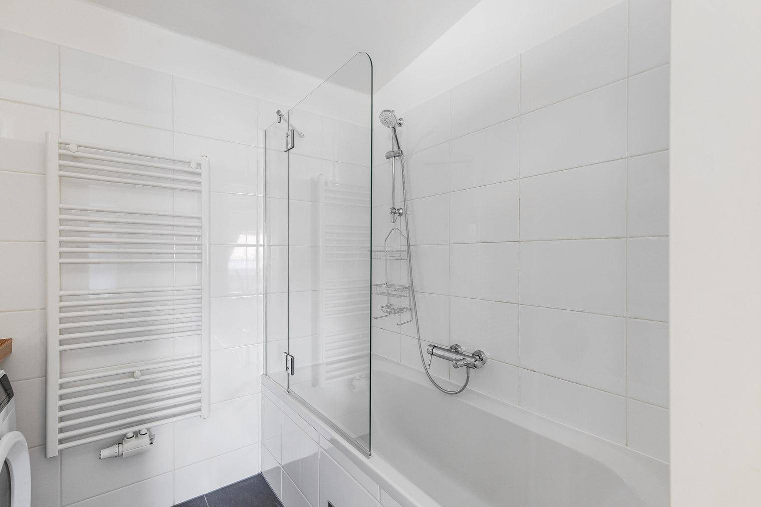 Helle Badewanne mit Glasabtrennung und Duschkopf in einem gefliesten Badezimmer.
