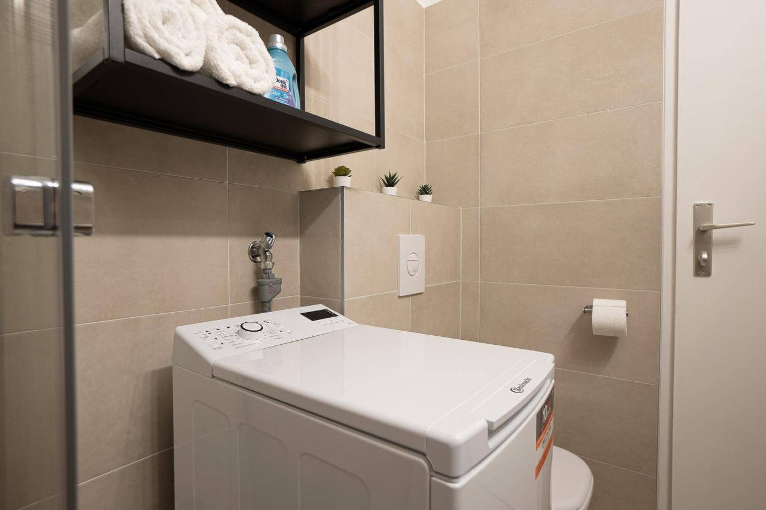 Badezimmer mit Waschmaschine, Regal und Wand-WC in neutralen Farbtönen, praktisch eingerichtet.