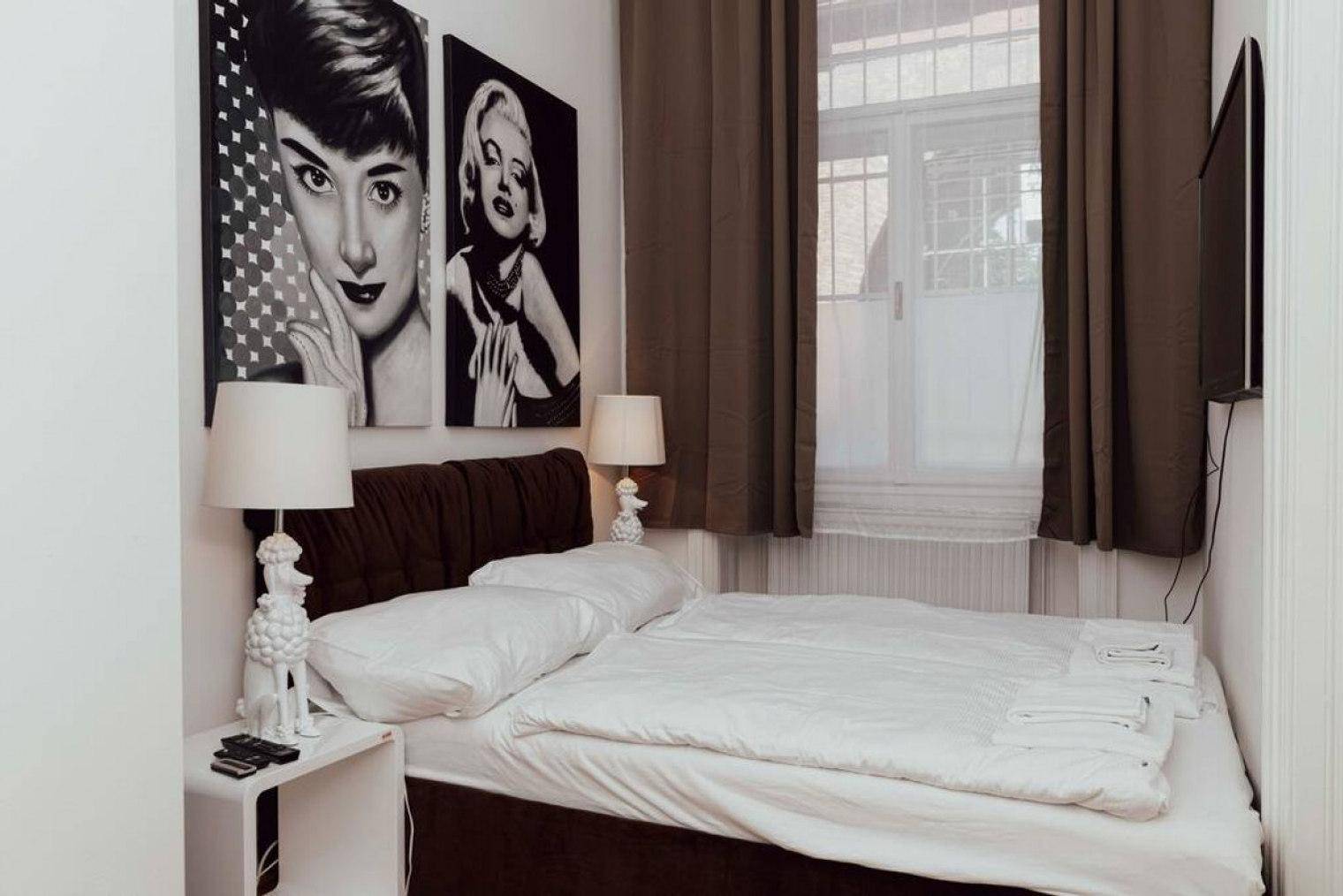 Schlafzimmer mit Doppelbett, Pop-Art-Bildern und dunklen Vorhängen vor dem Fenster.