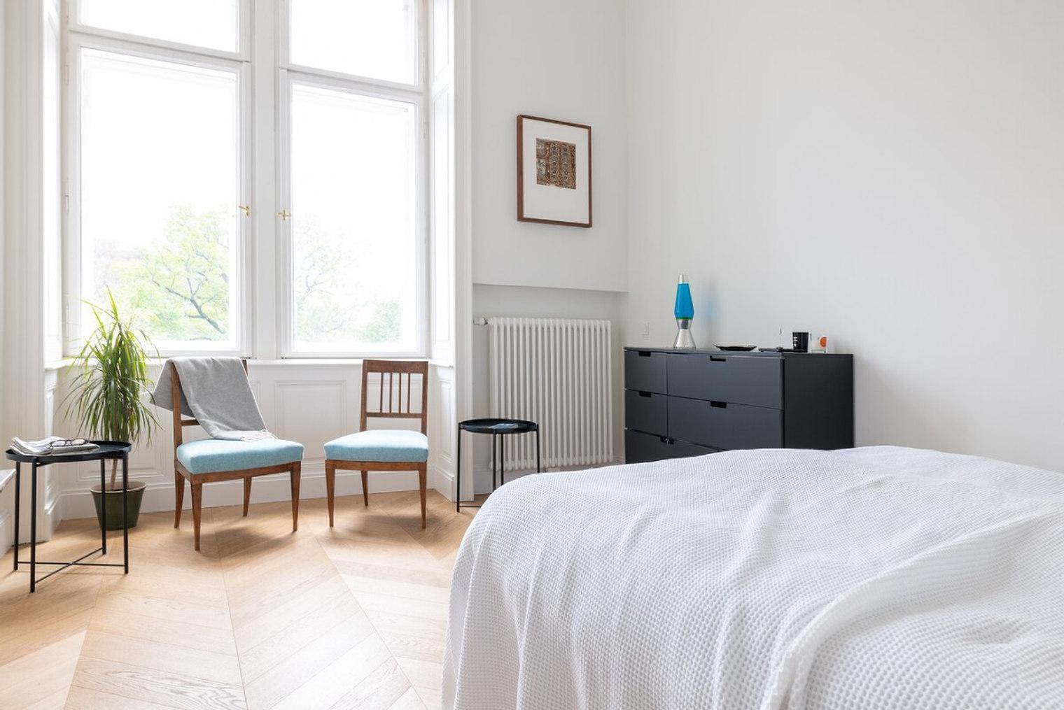 Helles Schlafzimmer mit großen Fenstern, Fischgrätparkett und einem bequemen Bett.