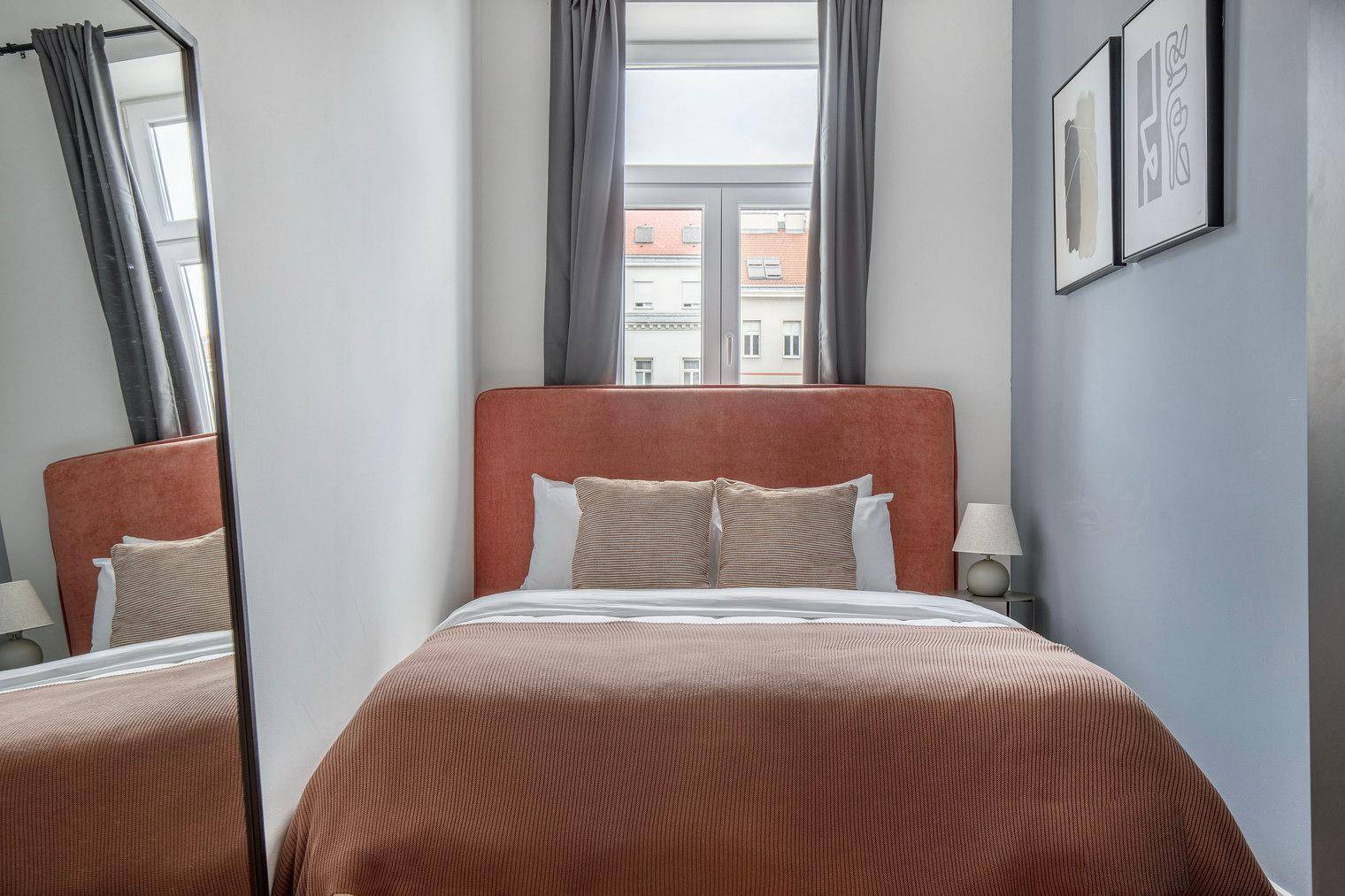 Helles Schlafzimmer mit Doppelbett, Spiegel und Blick auf die Stadt durch das Fenster.