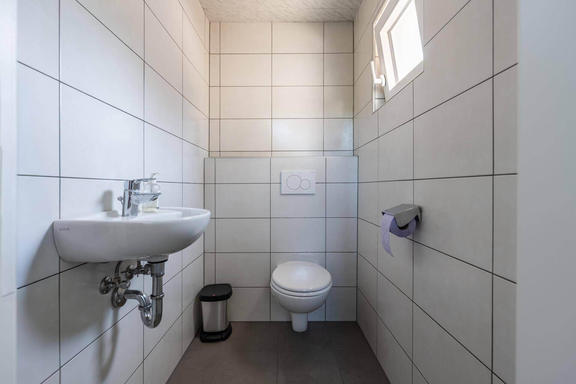 Modern gefliestes Gäste-WC mit Hänge-WC, Waschbecken und Fenster für Tageslicht, sauber und funktional.