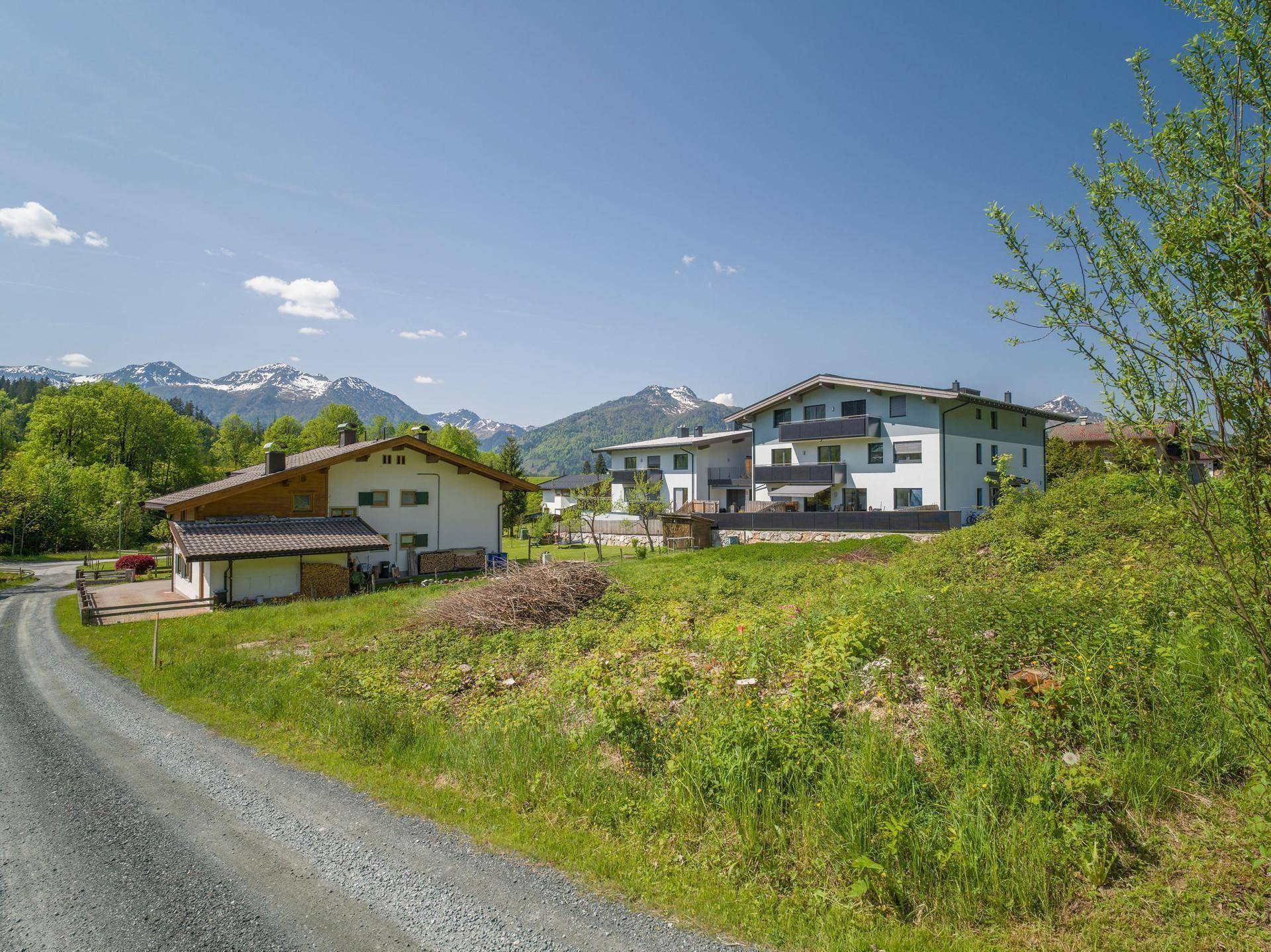 Ländliche Umgebung mit Wohngebäuden, einem Feldweg und majestätischen Bergen im Hintergrund.