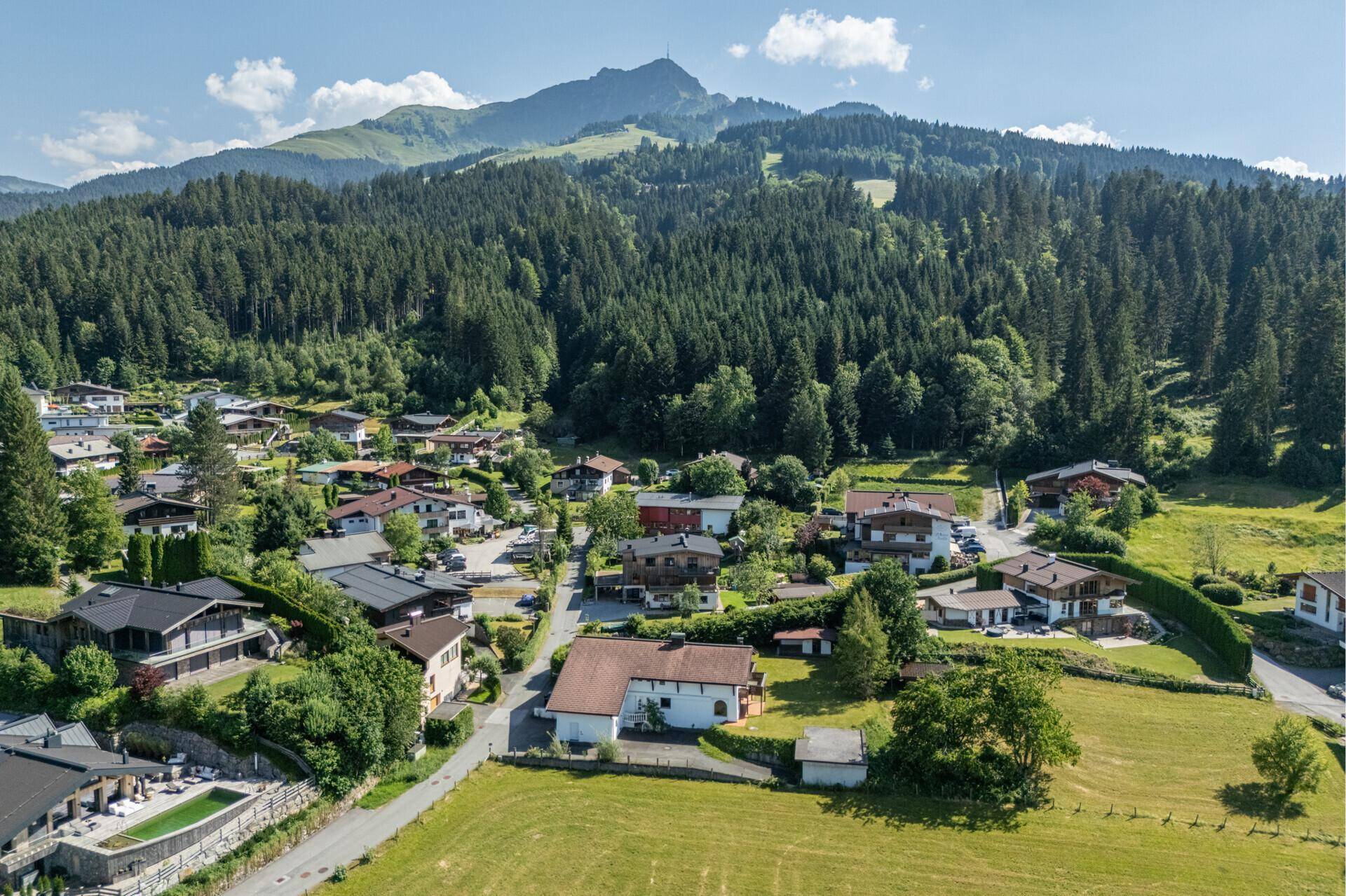 Luftaufnahme einer idyllischen Wohngegend, umgeben von dichten Wäldern und majestätischen Bergen unter blauem Himmel.